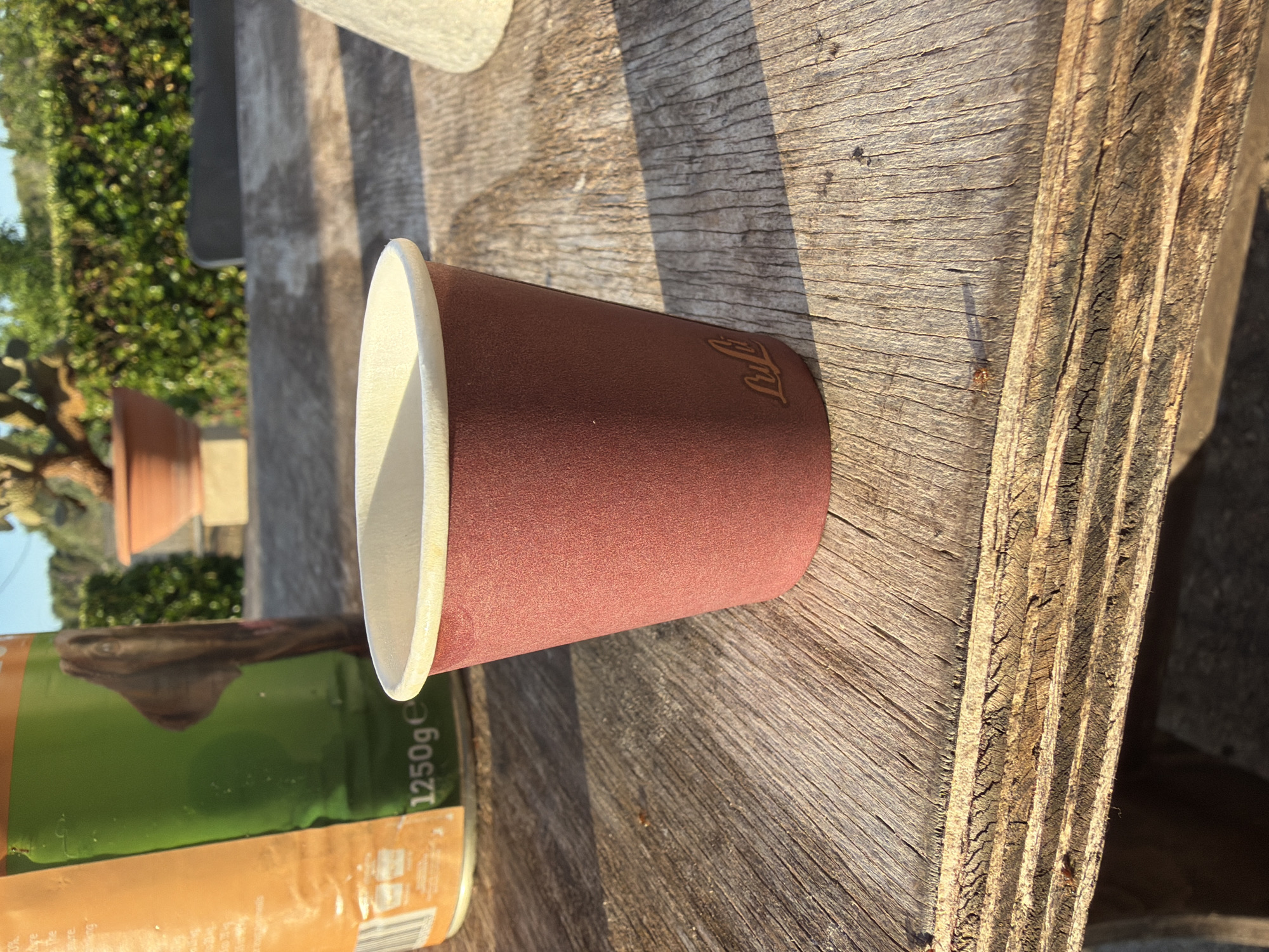 A close-up shot focuses on a small, maroon paper coffee cup with a beige rim, positioned on a rustic wooden table. The cup has a subtle embossed logo that appears to be the word "Luli". To the left of the cup, a portion of a large green and orange cylindrical object, possibly a can or container, is visible. The background suggests an outdoor setting with dappled sunlight creating shadows on the table. In the further background, there is greenery, including a hedge and what looks like a potted plant. The overall impression is that of an outdoor setting, potentially in a garden or patio area, with a casual arrangement of objects.