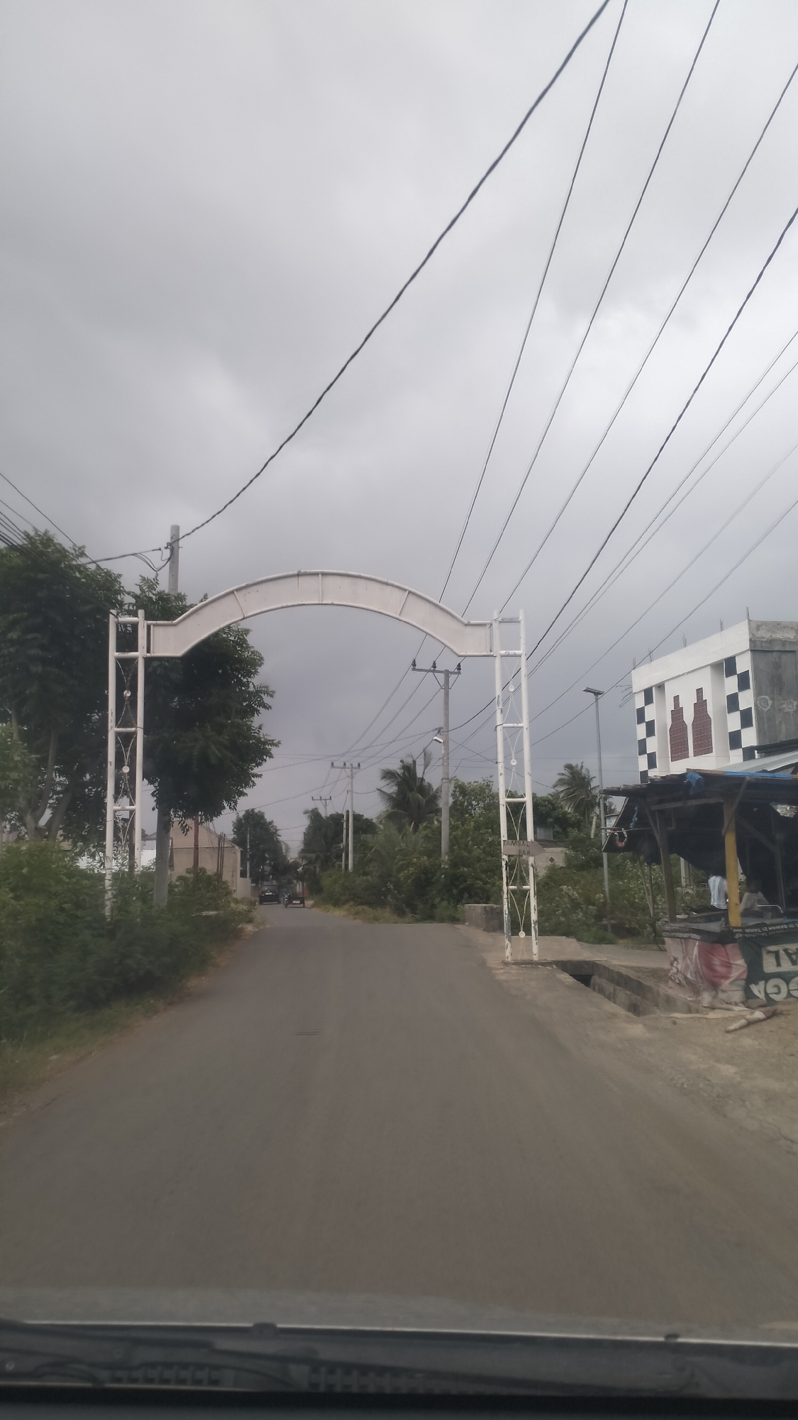 The image captures a street view in Miruk, Indonesia, dominated by a white archway crossing the road. The archway has decorative lattice work on its vertical supports. Overhead, a dense network of electrical wires crisscrosses the sky, which is overcast. To the right of the road, there is a building with a checkered facade and two prominent bottle-shaped designs. Adjacent to this building is a stall or small shop with a dark awning. The road itself is paved and appears to be a two-lane street, with greenery lining both sides. In the distance, two vehicles are visible on the road. The perspective suggests the photo was taken from inside a vehicle, looking forward.