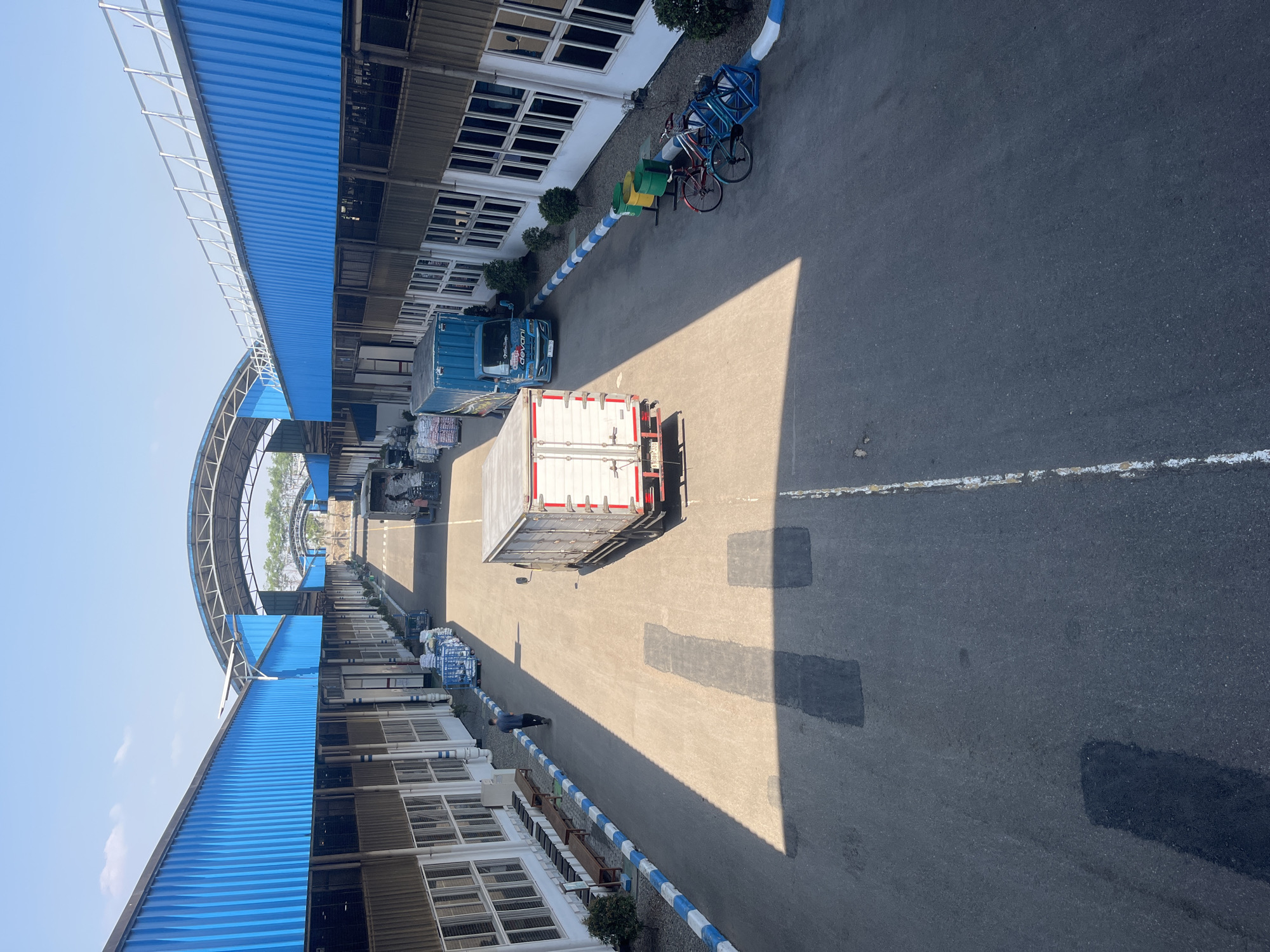 This image shows a wide paved roadway between two long, low-rise industrial buildings with arched, blue-paneled roofs. The buildings have white facades with numerous large windows and are bordered by a blue and white striped curb. Benches are positioned along the left building, and a single person walks down the road. On the right side of the road, there are several bicycles, trash receptacles, and a parked blue truck. Further down the road, a white truck is in motion, and more vehicles and trolleys loaded with goods are visible. The scene is illuminated by bright sunlight, casting distinct shadows on the pavement, indicating a clear day. The environment suggests an industrial or logistics hub. The location is identified as Pelang, Indonesia.