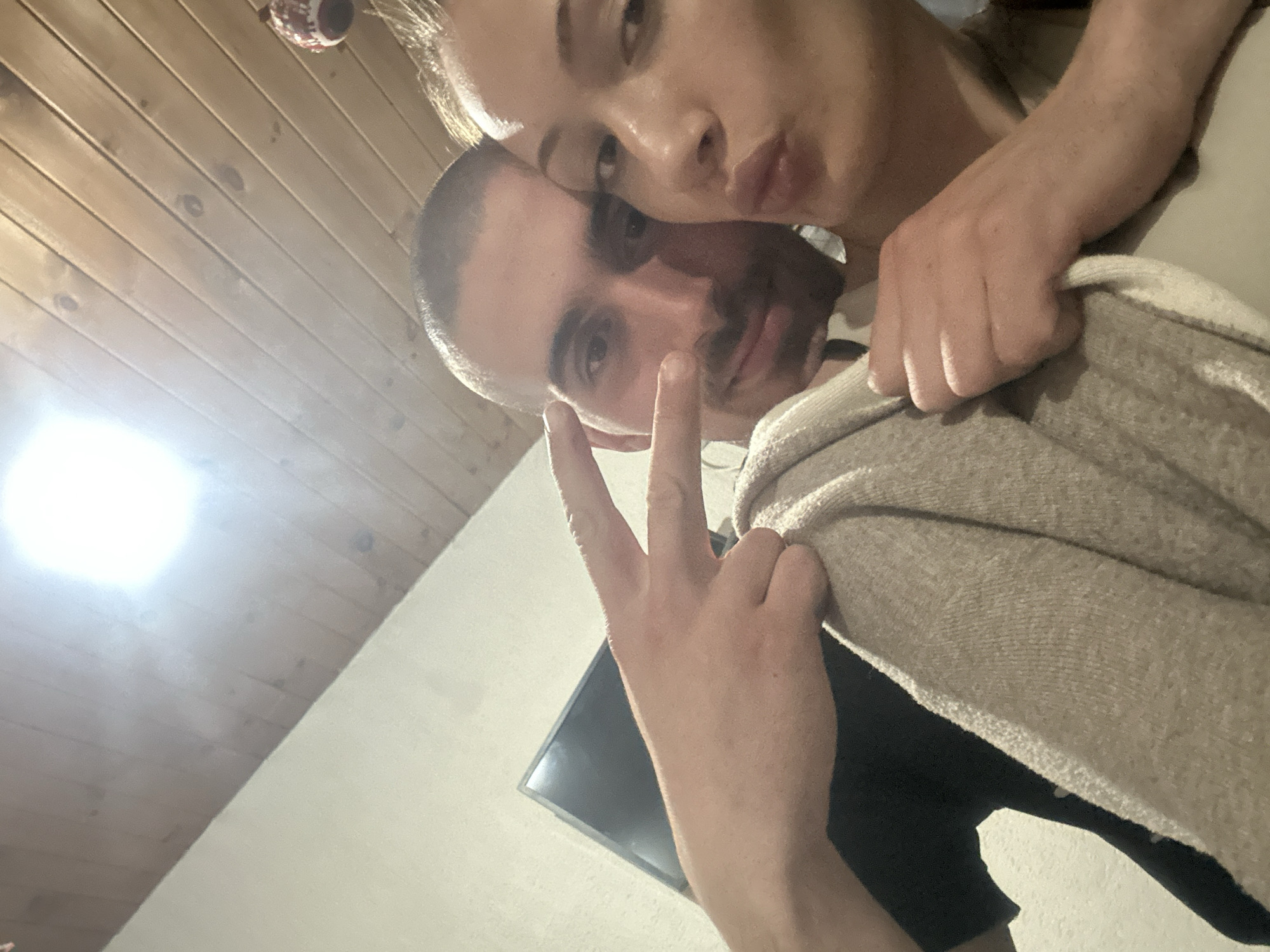 The image shows two people posing for a selfie in an indoor setting with a wooden ceiling and a light-colored wall. A television screen is visible in the background. The man is making a peace sign. The location is unknown and cannot be identified as Burgas, Bulgaria.
