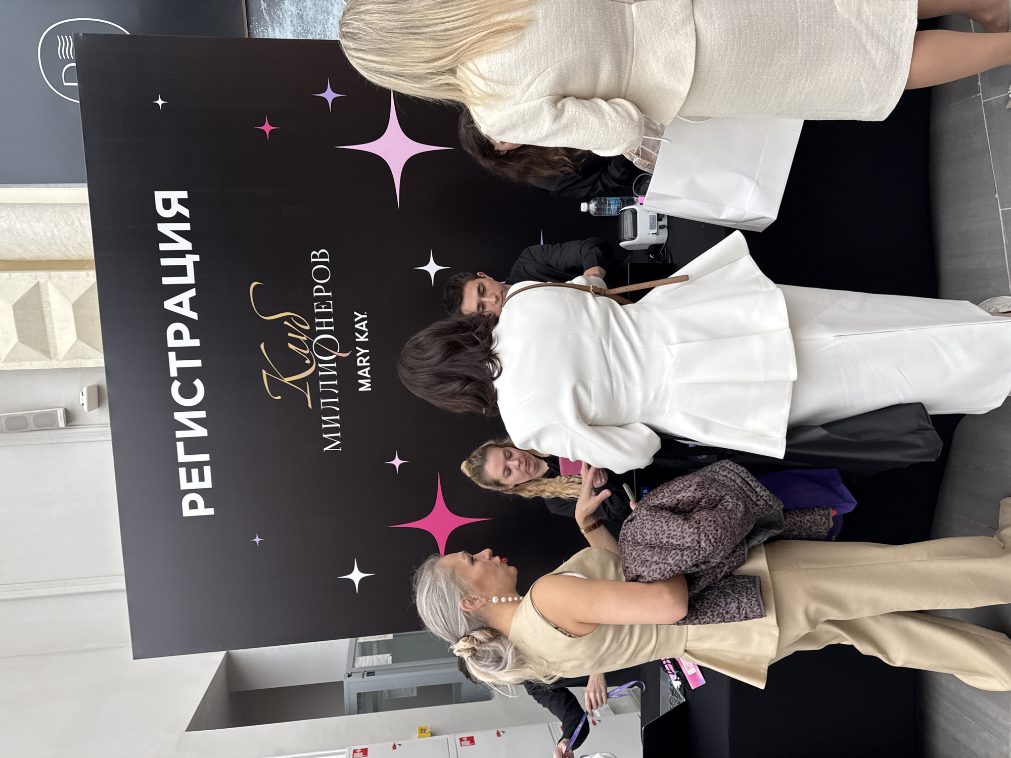 The image depicts a registration area for an event, likely a conference or promotional event. In the foreground, a woman with gray hair pulled back in a bun and wearing a beige suit is holding a leopard-print jacket and a purple bag. She is interacting with a woman in a white blazer who is looking down. Behind them, a man in a black shirt is working at a desk with a white device and a bottle of water. Another woman in a black shirt is also visible behind the desk. The backdrop is a large black banner with white text that reads "РЕГИСТРАЦИЯ" (Registration) and "КЛУБ МИЛЛИОНЕРОВ MARY KAY." (Millionaires Club Mary Kay.) There are also decorative pink and purple stars on the banner. The environment appears to be indoors, possibly a convention center or a large event hall, with concrete floors and a glimpse of a gray wall with structural elements at the top. The scene suggests an organized event is underway, with attendees checking in and staff assisting them. The overall impression is one of a professional gathering. The presence of the Mary Kay branding indicates a beauty or direct sales industry event. The specific location in Moscow, Russia, is not explicitly visible in the image itself, but the Cyrillic text on the banner confirms it is in Russia.