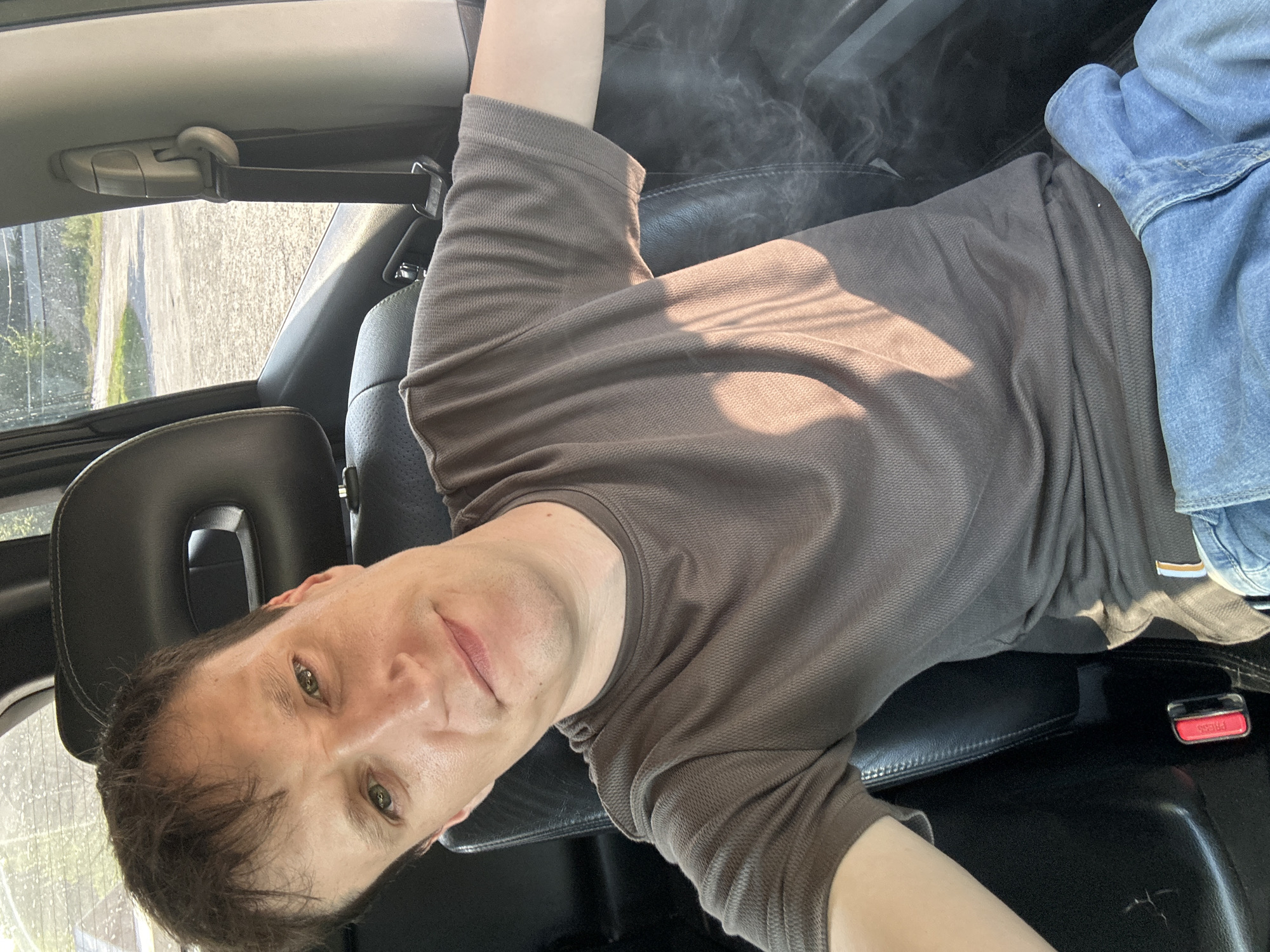 A medium shot captures a man seated in the driver's seat of a vehicle, looking directly at the camera. He has light skin, short brown hair, and green eyes. He is wearing a brown, short-sleeved, textured t-shirt and blue jeans. Sunlight casts shadows across his t-shirt. The interior of the car is visible, including a black leather seat, a seatbelt with a red buckle, and the headrest. Through the window, a dirt road and trees are visible, suggesting an outdoor location, possibly in Makiv, Ukraine. The background is slightly blurred.