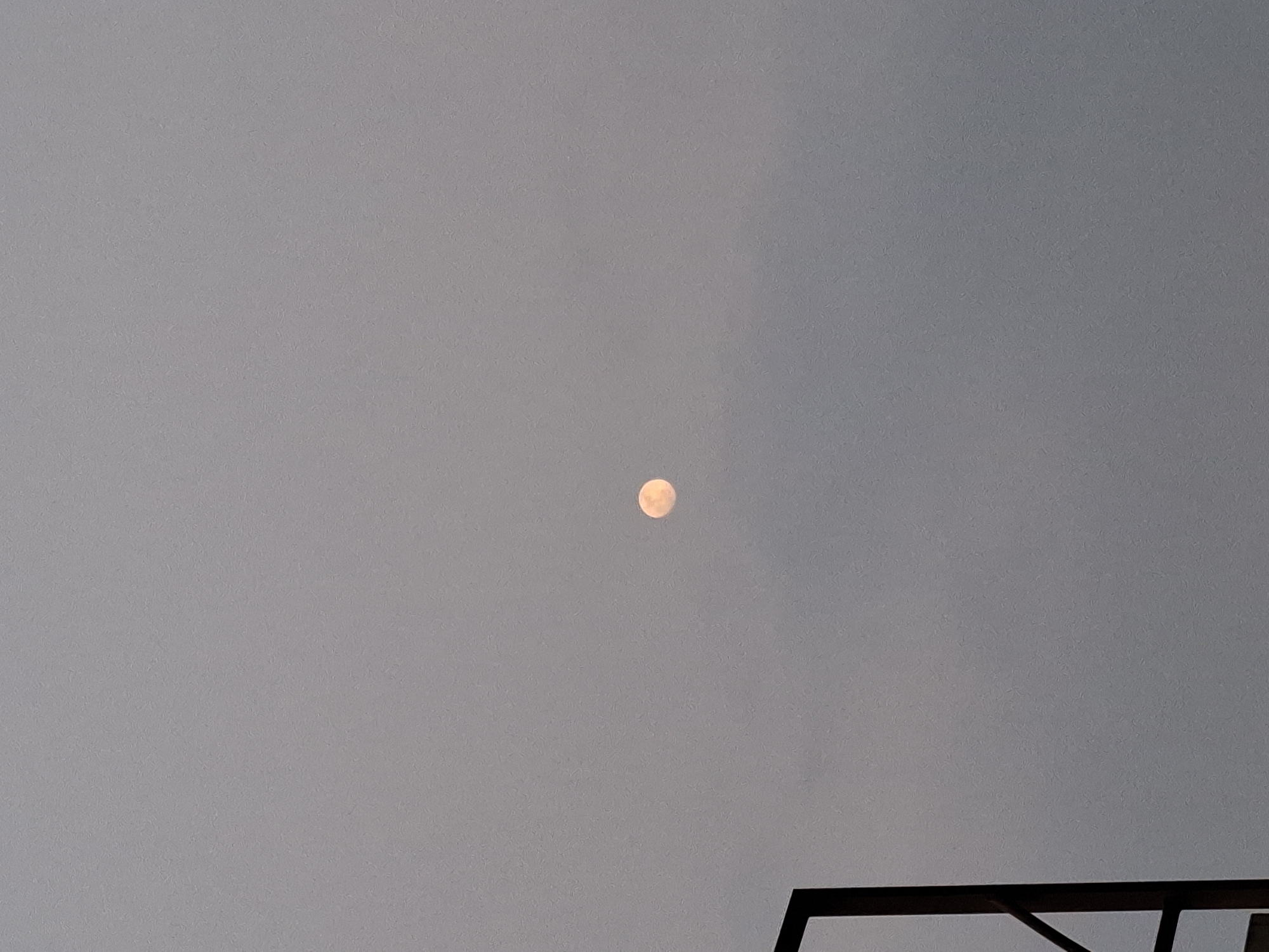 A full moon, appearing slightly orange or peach-colored, is visible in the overcast sky. To the left of the frame, a dark, angular structure, possibly part of a building or scaffolding, extends from the bottom edge upwards. The background is a uniform, muted gray due to the cloud cover. The scene suggests an outdoor view in Sampora, Indonesia, during twilight or under heavy cloud conditions.