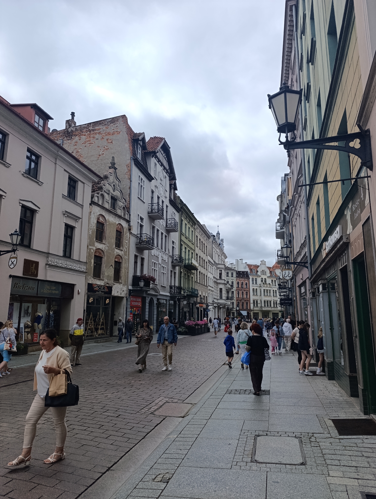 The image depicts a view of a busy pedestrian street in Toruń, Poland, likely Długa street, given the architecture and shops. The street is paved with cobblestones and lined with colorful, historic buildings on both sides. Many of these buildings feature ornate facades and balconies, indicative of European architectural styles.
On the left side of the street, a woman in a cream-colored cardigan and light-colored pants walks towards the viewer, carrying a dark blue and beige shopping bag. She is wearing sandals and appears to be casually dressed. In the background, various shops with glass storefronts are visible, with some displaying mannequins. A black lamppost with a decorative bracket is mounted on the wall of one of the buildings.
On the right side of the street, more shops line the sidewalk, with people browsing or walking by. The buildings on this side are also multi-story and have varied colored facades. A prominent black lantern-style lamp is mounted on a bracket extending from one of the buildings.
The street is populated with numerous people walking in both directions, suggesting a lively commercial and tourist area. The people are dressed in a variety of casual attire, including t-shirts, jeans, dresses, and jackets. Some are walking alone, while others are in small groups.
The sky is overcast and appears to be cloudy, contributing to a diffused light throughout the scene. The overall atmosphere suggests a typical day in a European city, with daily life and commerce unfolding on a historic street.