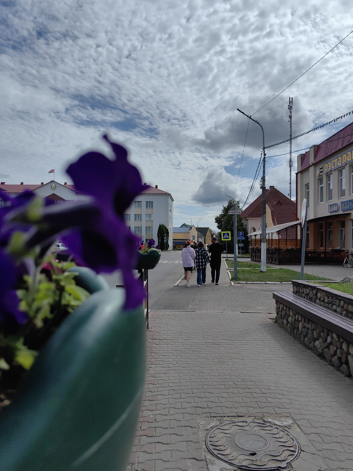 A street scene in Pastavy, Belarus, captures three individuals walking away from the camera on a paved sidewalk. The group consists of two women and one man, all dressed casually. To the right of the sidewalk is a stone-and-wood bench. In the foreground, out of focus, is a green planter with purple flowers. The background reveals buildings, including one with signage that appears to be in Cyrillic script and reads "РЕСТОРАН." A red-roofed building is also visible. Overhead, the sky is filled with white, puffy clouds against a blue backdrop. Streetlights and utility poles line the street, with a cell tower present. A pedestrian crossing sign is visible near the sidewalk. A manhole cover is set into the pavement.