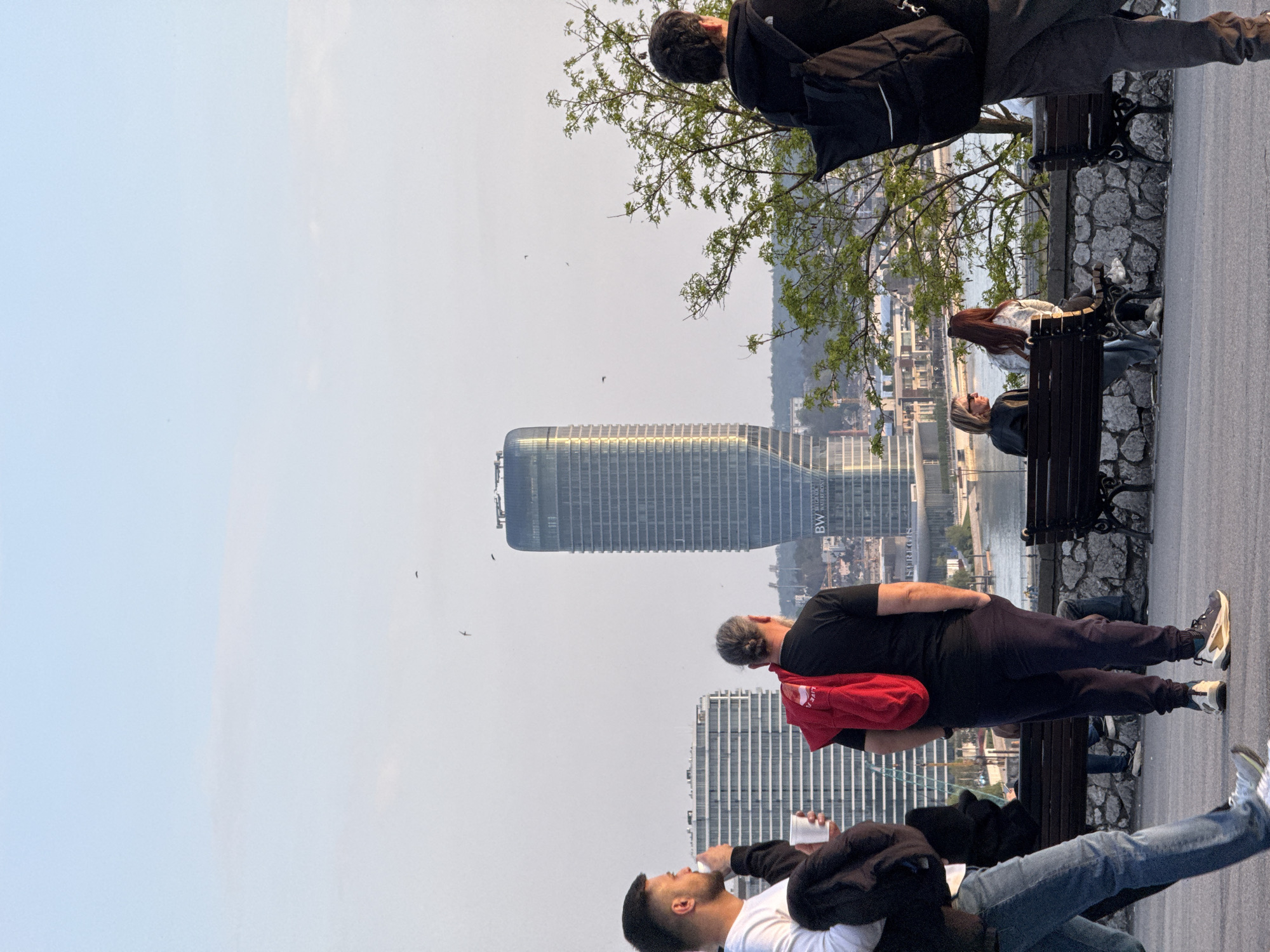 The image shows an elevated perspective of a cityscape, likely Belgrade, Serbia, evident from the modern architecture. A prominent skyscraper with a curved top dominates the view. People are visible in the foreground, some standing, some sitting on benches. A man in a black jacket is standing near a tree. A man in a black shirt and a red backpack is looking at the cityscape. Another man is drinking from a cup. The sky is overcast and hazy, with some birds in the air.