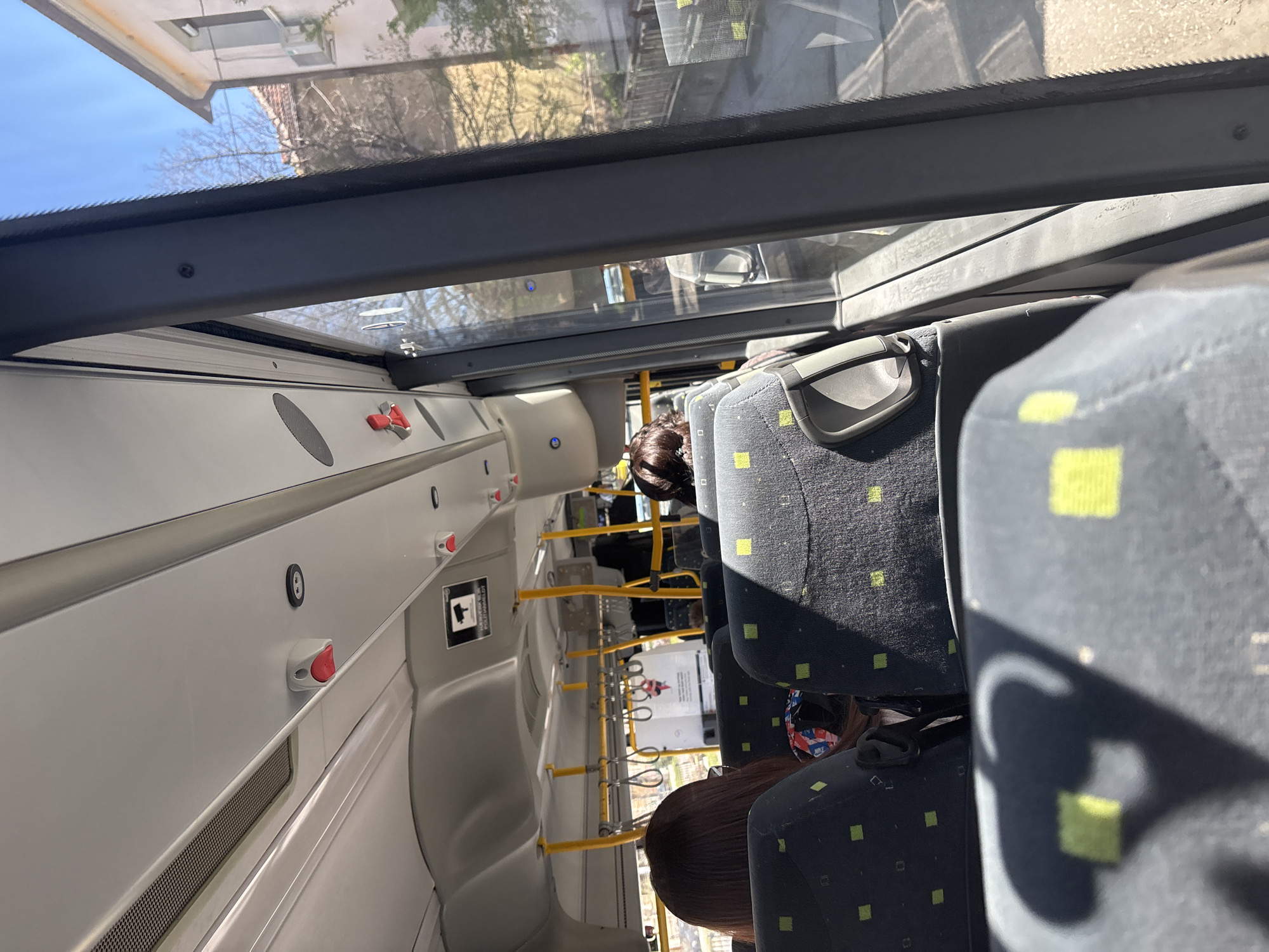 The image depicts the interior of a bus in Plovdiv, Bulgaria. Passengers are seated in patterned, green-and-gray seats. A row of yellow handrails runs along the aisle. The bus interior is white with red emergency stop buttons. The exterior is visible through the windows.