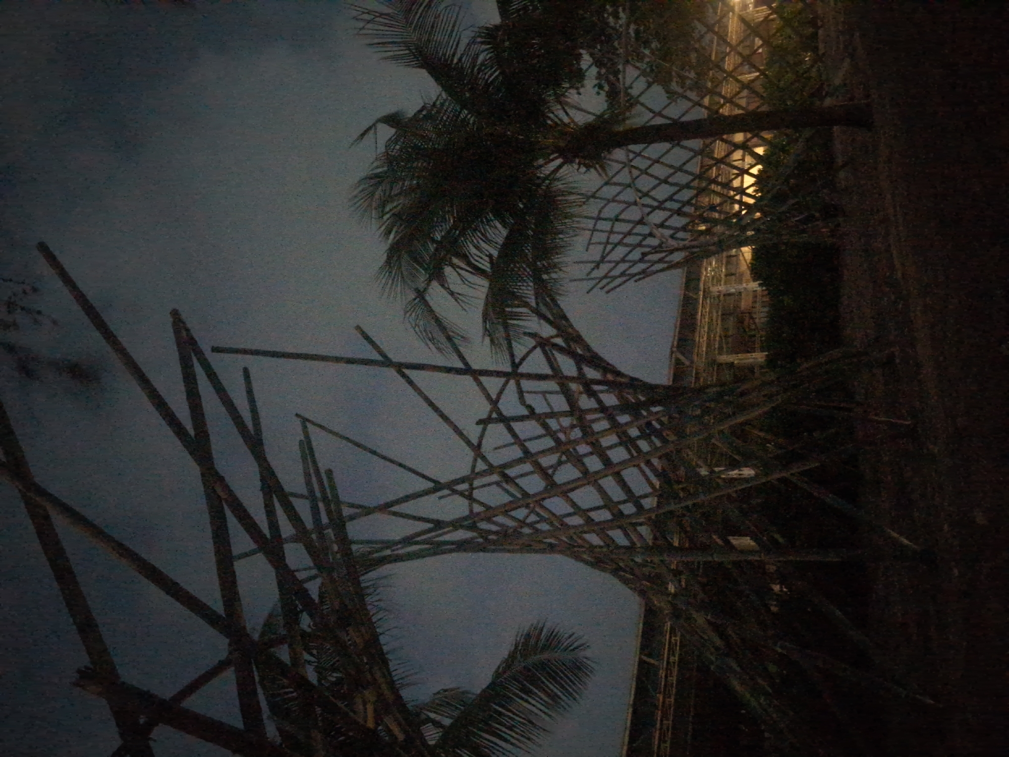 The image depicts a low-angle shot of a structure made of bamboo poles and woven lattice, likely a temporary construction or decorative element. Palm trees are partially visible, framing the structure against a dusky sky. The image appears to be taken at night or in dim light. The location is likely in Phú Quốc, Vietnam, based on the palm trees and architectural style. A multistory structure is partially visible. The photograph's orientation is rotated.