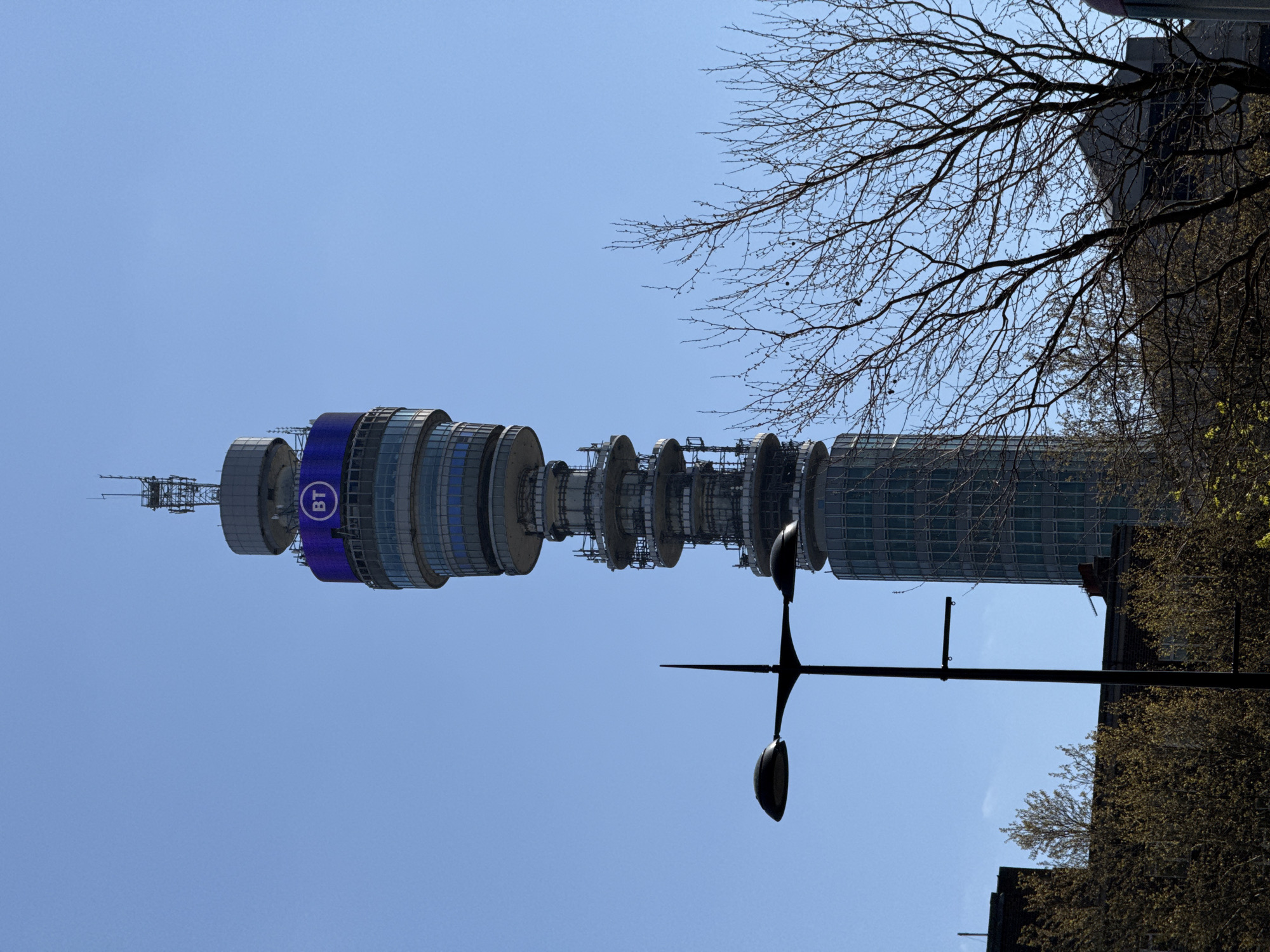 The image shows the BT Tower in London, United Kingdom, from a low angle. The tower is cylindrical and consists of multiple sections. The BT logo is visible on a blue band around one section. The sky is clear and blue. A lamppost with two lamps and a spike is in the foreground. Bare tree branches and the side of a building are visible on the right. Trees with budding leaves are also present.