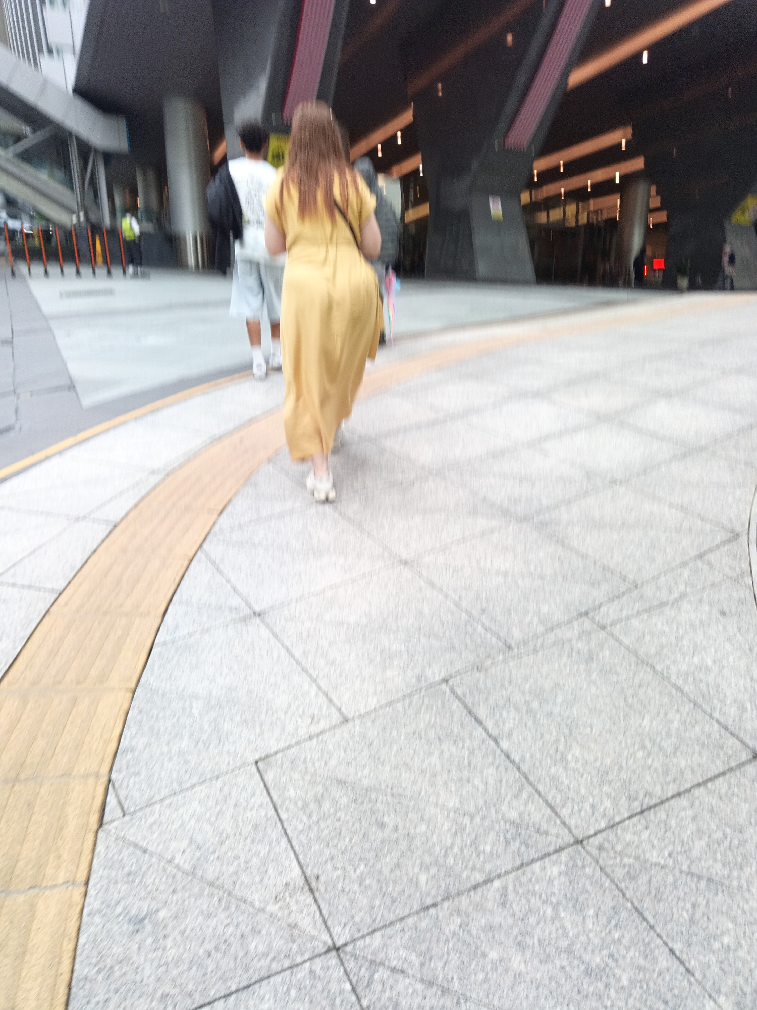 A long shot captures a woman in a long yellow dress walking away from the camera on a patterned sidewalk in Kuala Lumpur, Malaysia. She is followed by a man in a white t-shirt and light-colored shorts, who is carrying a black bag over his shoulder. The background features a modern building with a dark, angled roof structure and pillars. The sidewalk has a textured yellow section that curves to the left of the frame.