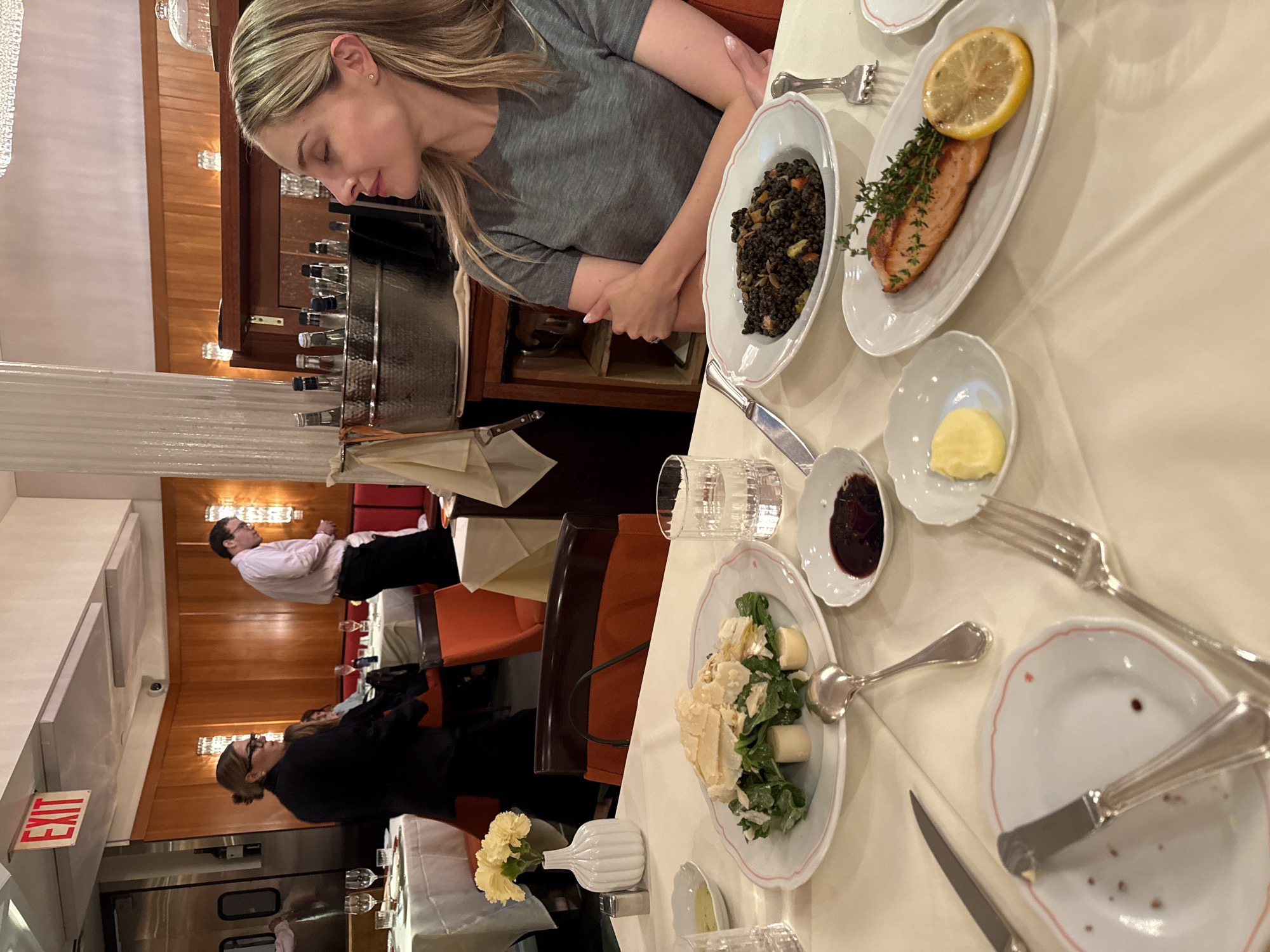 This image depicts a dinner scene inside a restaurant. A woman with blonde hair is seated at a table, her head turned away from the camera, with her arms crossed. In the foreground, a table is set with multiple plates of food, including a salad with shaved cheese and what appears to be white asparagus, a dish of black lentils with vegetables, and a piece of salmon with a lemon slice and herbs. There are also glasses of water, silverware, and small bowls of sauce and butter. In the background, two people are visible; one in a dark suit looking down, and another in a light-colored shirt, possibly a waiter. The restaurant interior features warm wood paneling, a prominent "EXIT" sign, and ambient lighting. The scene is captured from a slightly elevated perspective, looking down onto the table. The setting suggests a formal or upscale dining establishment.