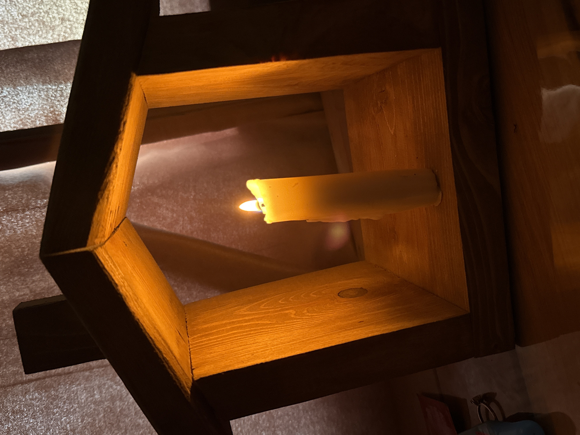 A burning candle is fixed to a wooden structure. The structure is a series of nested squares of wood. The wood grain is visible. The candle is cylindrical and pale yellow with a bright orange flame. The background is out of focus, appearing to be draped fabric. Based on the prompt request for the location of Tyulyuk, Russia, the assumption is that this image was taken in that location. There are no people visible.