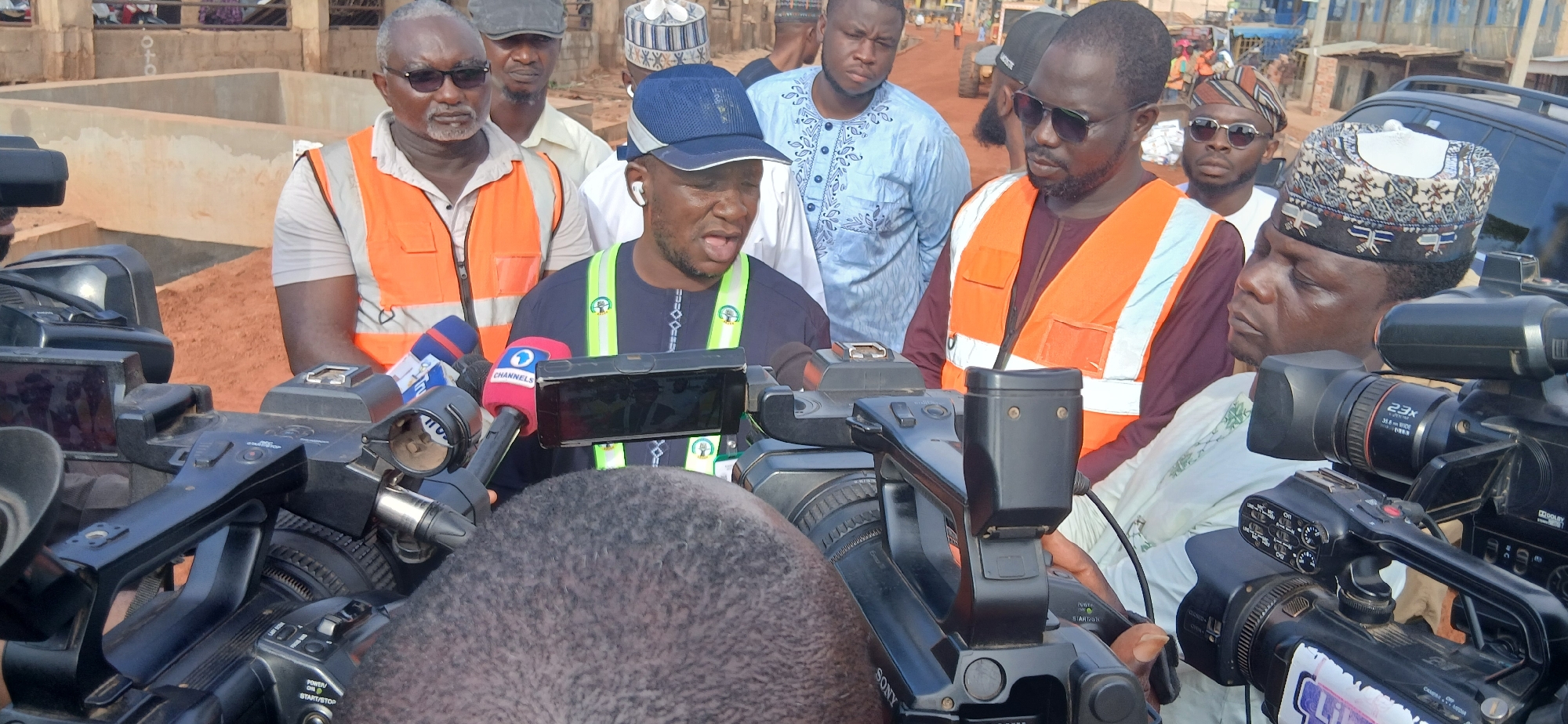 In Zaria, Nigeria, a group of men are gathered outdoors, with a focus on two individuals speaking to a cluster of microphones from multiple news organizations. One man, wearing a blue baseball cap and a dark blue, embroidered garment with reflective yellow trim, appears to be addressing the press. He is surrounded by microphones, including one with the "Channels" logo. The other prominent individual, to his left, is an older man with glasses, wearing an orange safety vest over a light-colored shirt. He is looking towards the speaker. Other men in the background are also present, some wearing caps and vests, suggesting a work site or official gathering. The environment appears to be dusty with construction materials and some structures visible in the background. Several professional video cameras are positioned in the foreground, indicating a media event.