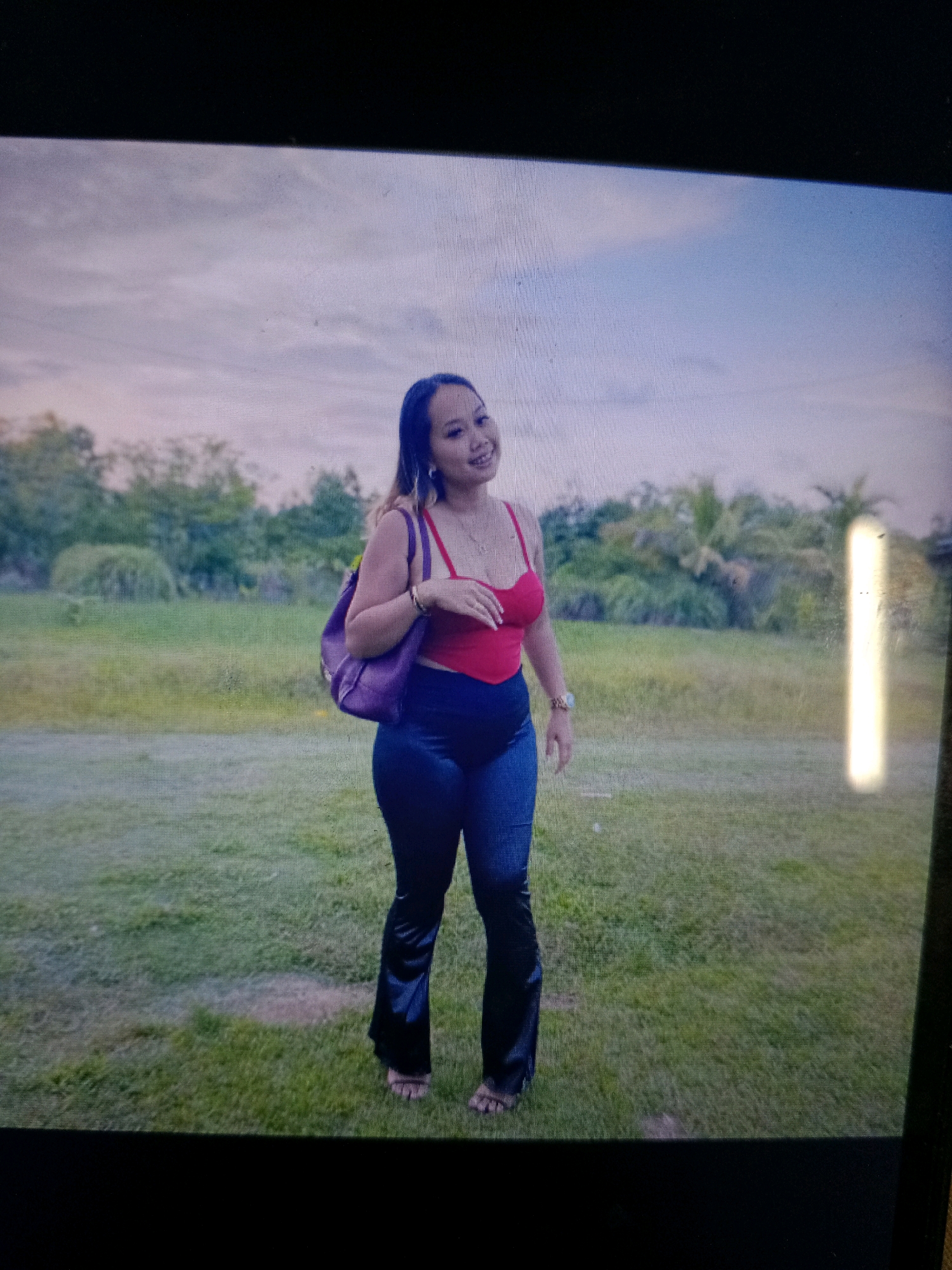 In the city of Noord, Suriname, a woman stands outdoors in a grassy field. She is wearing a red tank top, dark flared pants, and flip-flops. A purple backpack is slung over her left shoulder. The background features a line of trees and vegetation under a cloudy sky. The lighting suggests it is daytime.