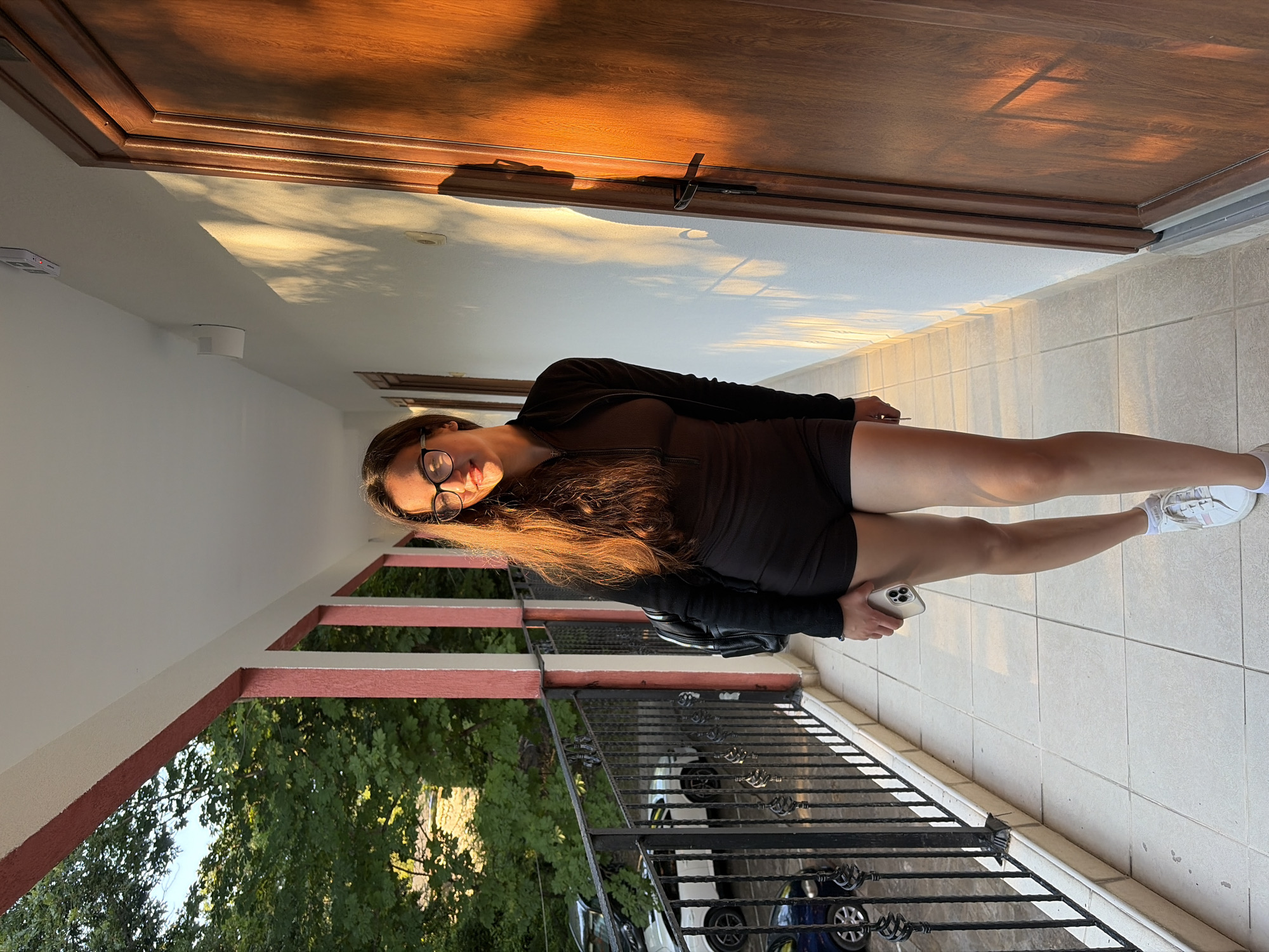 A young woman stands on a balcony in Kiten, Bulgaria, bathed in the warm, golden light of the setting sun. She is wearing a dark, fitted athletic outfit consisting of a long-sleeved zip-up top and shorts, along with white athletic shoes and glasses. Her long, brown hair is down, and she holds a smartphone in her right hand. She has a backpack slung over her left shoulder. The balcony has a tiled floor and a railing overlooking a parking area with several cars, including a dark blue vehicle and a white one. Behind her, there are doorways leading into a building, and trees can be seen in the background, adding a touch of nature to the urban setting. The sunlight casts long shadows across the white wall of the building, creating a dramatic visual effect.