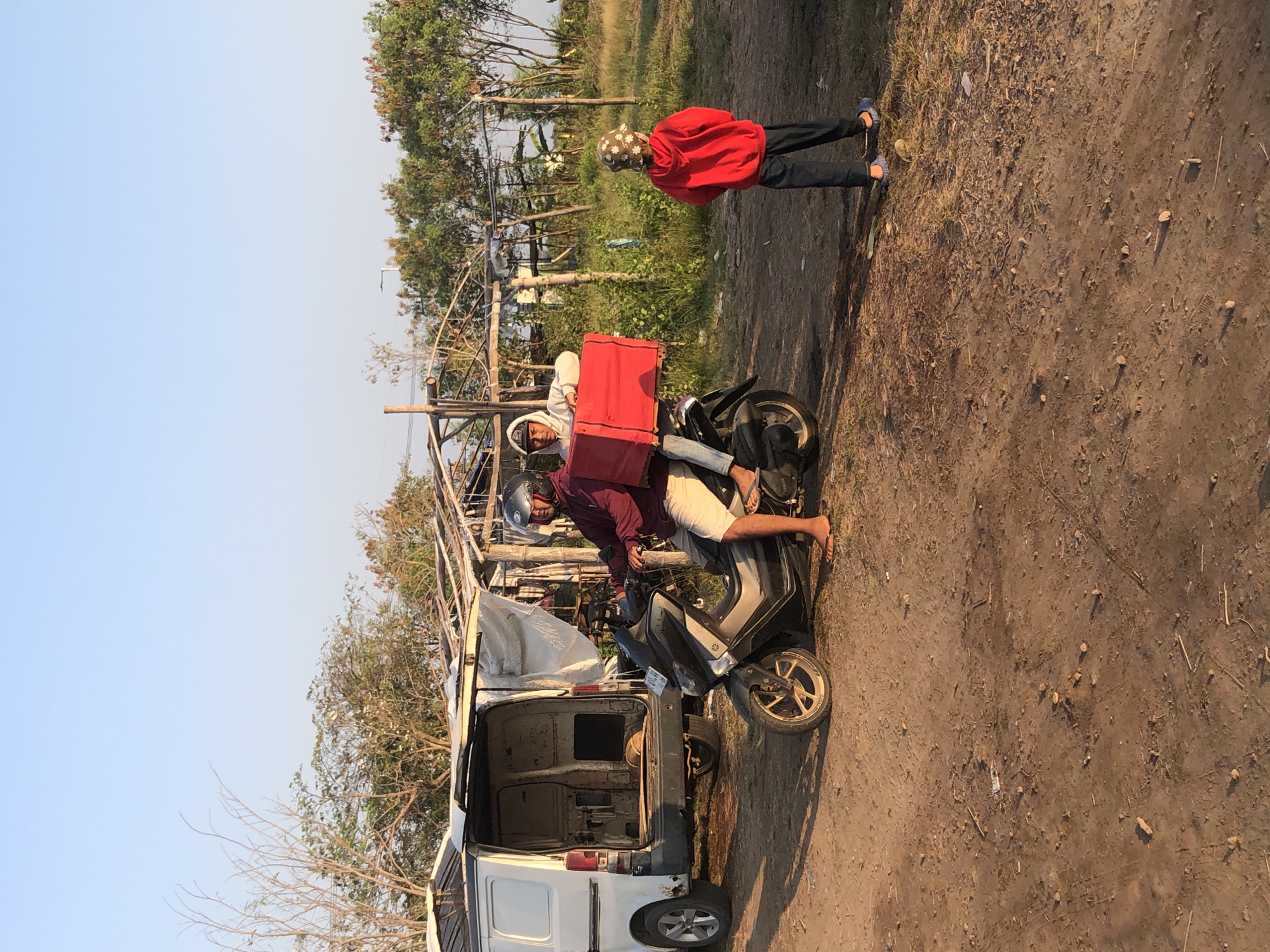 In Gempolsari, Indonesia, a snapshot captures a rural or semi-rural setting with a clear blue sky. The foreground is dominated by a dirt ground with scattered small stones and debris. To the left, the rear of a white van, its doors open, is visible. Beside it, a dark-colored motorcycle is parked. A man wearing a maroon jacket, white shorts, and a helmet is seated on the motorcycle, his right foot resting on the ground. He is facing towards the right. Behind him, another person, wearing a white hooded jacket and jeans, is seated on the motorcycle, holding onto a large red box secured to the rear. To the right of the motorcycle, a child stands facing away from the viewer, wearing a red hooded jacket, dark pants, and a patterned hat. The background features overgrown vegetation, trees, and a simple open-sided structure with a roof made of corrugated metal or tarpaulin, supported by wooden poles. A power line is visible in the distance. The scene appears to be taken during daylight, possibly late afternoon, given the warm lighting.