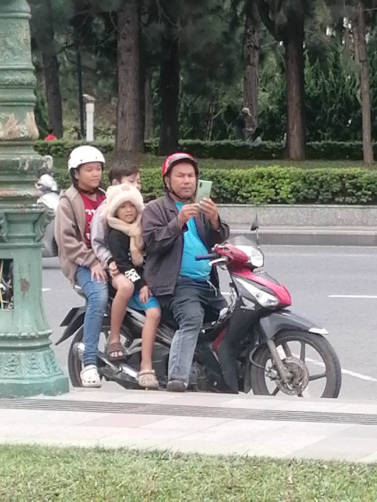 In Da Lat, Vietnam, a man wearing a red helmet and a blue shirt is operating a motorcycle, holding up a smartphone to take a picture. Behind him, a child in a fluffy white hat and scarf is seated between two other children. The child in front is wearing a white helmet and a brown jacket, while the child behind the driver wears a light-colored jacket. The motorcycle is black and red, and they are on a paved road with a pedestrian walkway and grass to the side, bordered by trees and bushes.