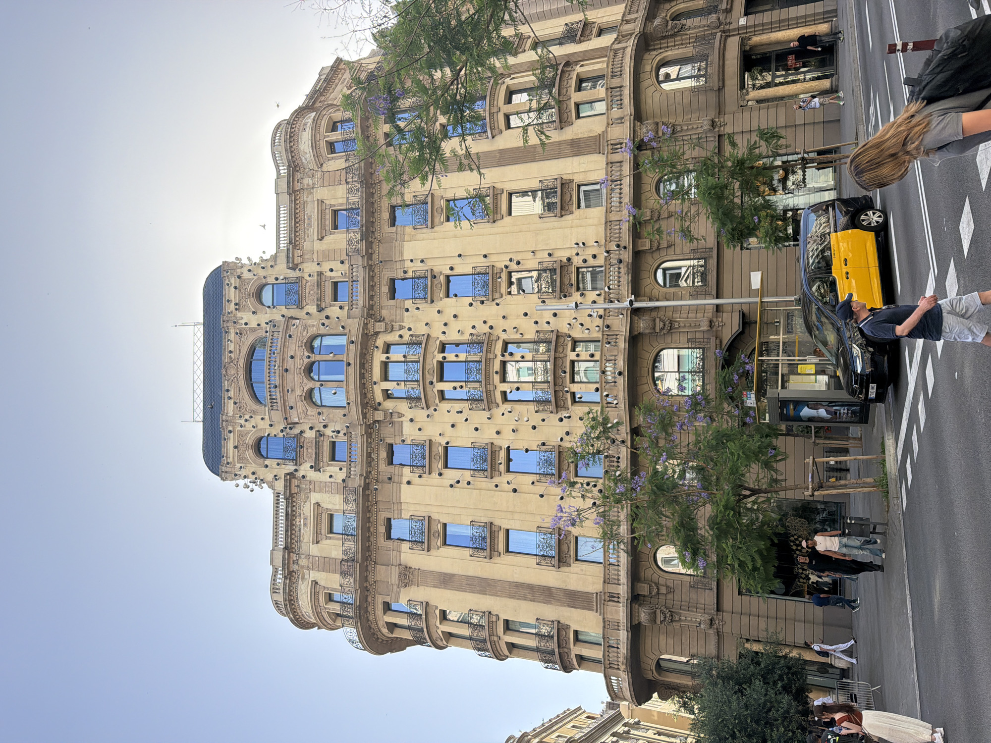 The image captures a street-level view of a multi-story, ornate building facade in Barcelona, Spain, likely on a sunny day with a clear sky. The building, characterized by its light brown stone and neoclassical architectural style, features numerous windows with blue panes, many adorned with intricate wrought-iron balconies. Several round, dark decorative elements are affixed to the facade, and trees with delicate purple blossoms are visible in front of and beside the structure. A yellow and black taxi is parked on the street in front of the building, alongside a streetcar or bus stop shelter. Pedestrians are seen walking on the sidewalk and crossing the street, some appearing to be tourists. The overall scene depicts a vibrant urban environment with a blend of historic architecture and contemporary city life.
