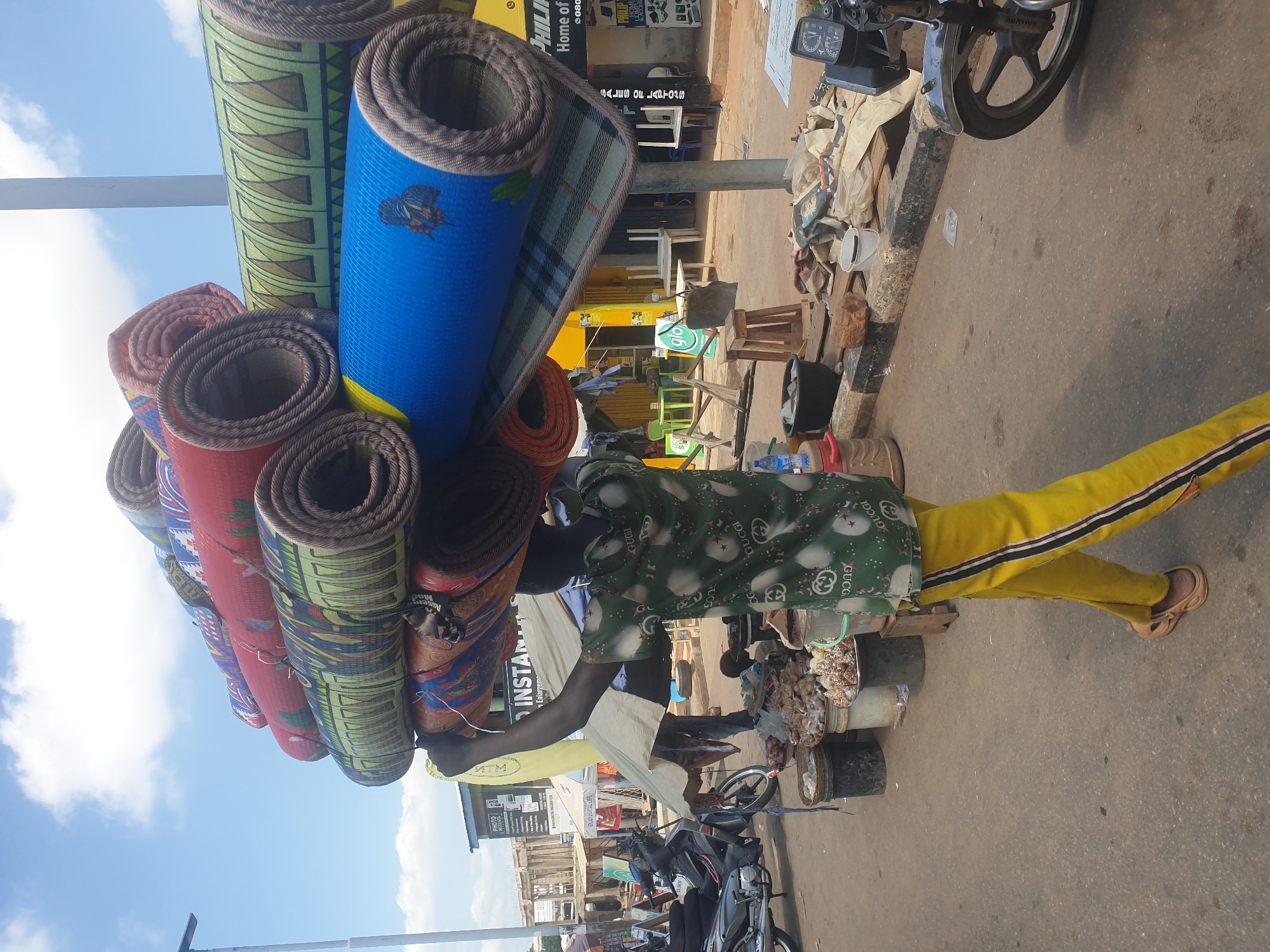 An African man in Lokoja, Nigeria, is carrying a bundle of rolled mats on his head along a street. The man is wearing yellow pants with black stripes, a green shirt, and brown sandals. The mats are of various colors and patterns. To the right of the man is a motorcycle parked on the side of the road with various items in baskets and buckets attached to the side. Behind the man, there are various shops and businesses. The sky is visible in the upper left corner. The street appears to be paved.