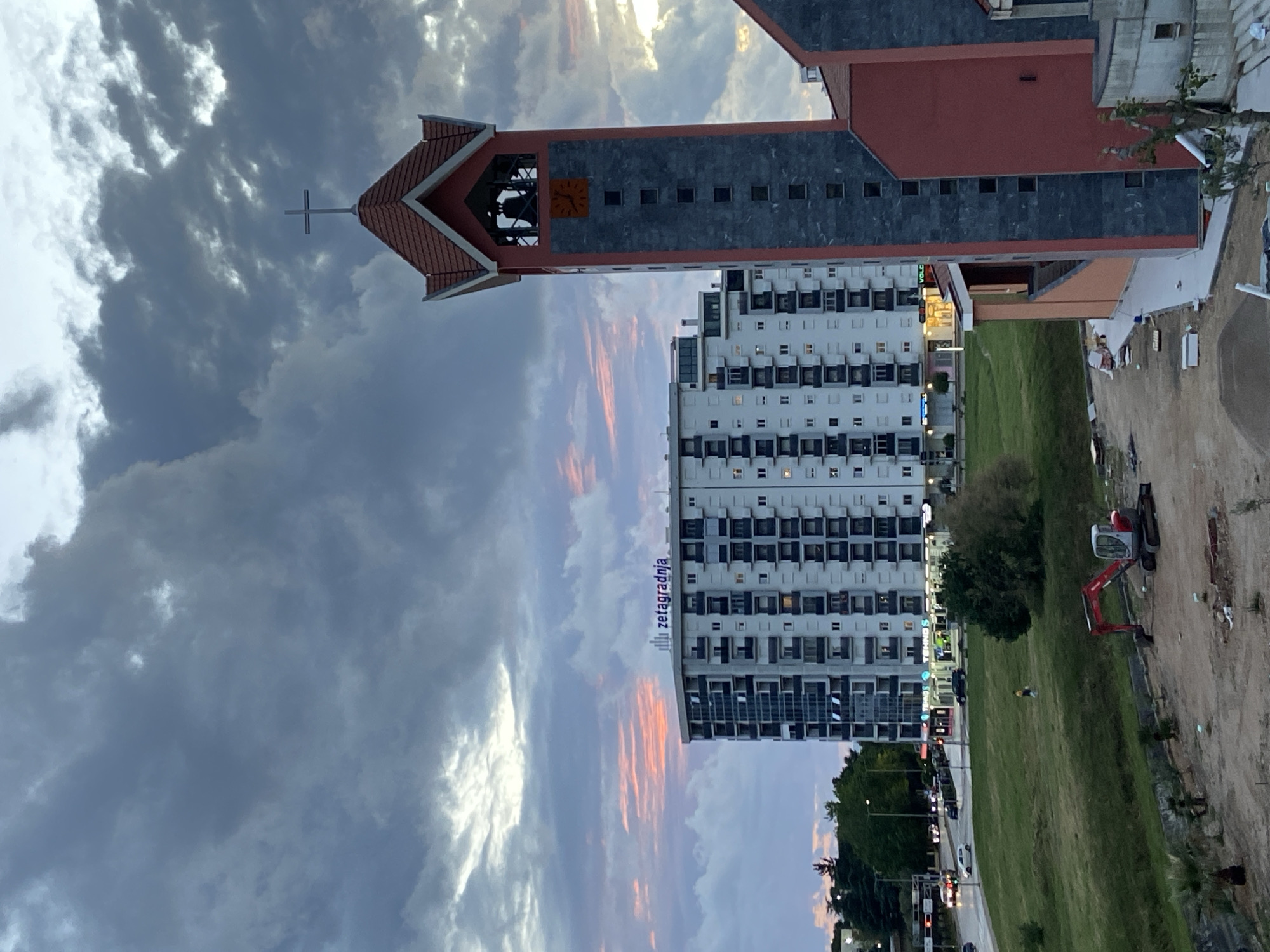 The image shows a view in Bar, Montenegro. The church tower is prominent, with a cross at the top. Next to it, there is a tall, modern building with the word "zetagradnja" on its side. Between them is a green strip of grass with street lights and traffic. On the right side of the scene, there is an area under construction with a small excavator. The sky is dramatic with cloudy patterns, including some pink hues from the setting sun.