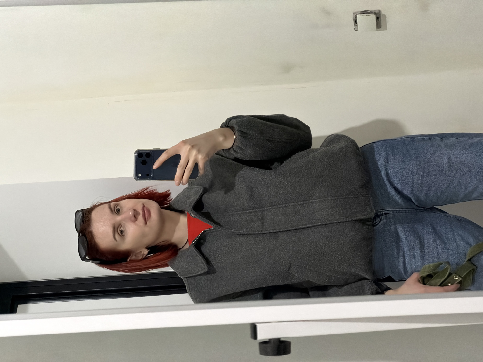 A woman stands in a white-walled room, taking a selfie with her mobile phone. She is wearing blue jeans, a red top, and a dark gray jacket. Sunglasses are perched on her head. Her hair is reddish-brown. She is holding a green strap in her right hand. The location is potentially within the city of Chaikovsky, Russia; however, there is no specific evidence within the image to confirm the precise location or city. The room is sparely furnished, with a black frame visible in the lower left and a metal bracket near the ceiling. The floor and additional elements are obscured.