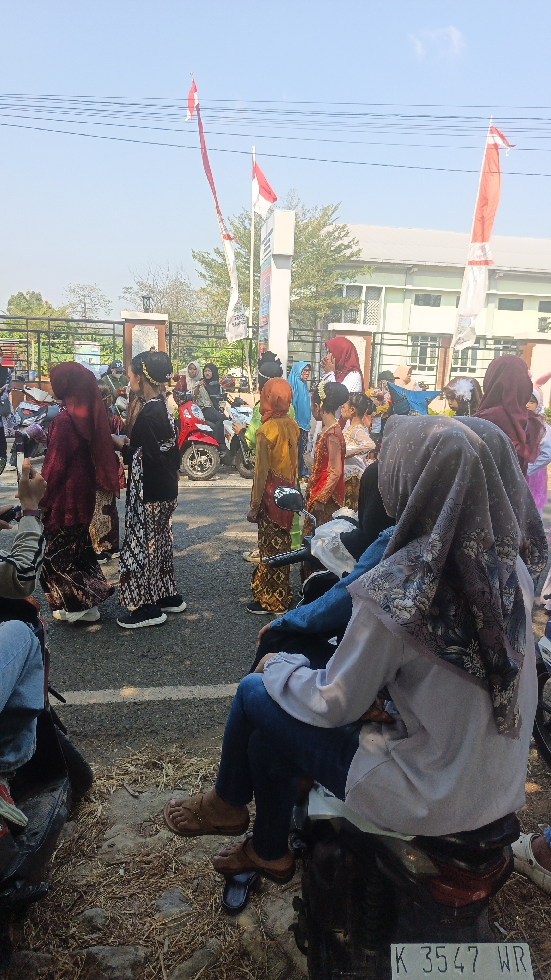 In an outdoor scene with a clear blue sky, a group of people, many of whom are children dressed in traditional attire with elaborate hairstyles, are gathered on a paved road. Indonesian flags are prominently displayed, with one flag at the top of a tall pole in the background and another larger flag on a pole to the right, both fluttering in the wind. A tall, white sign with text and graphics is visible behind some of the children. In the mid-ground, several motorcycles are parked. A building with a light-colored facade and green trim is visible in the background. In the foreground, two individuals are seated on a motorcycle, with their legs and lower torsos visible. One person is wearing blue jeans and brown sandals, while the other is wearing a light purple long-sleeved top and a patterned headscarf. The ground is covered with dry grass and debris. The license plate on the motorcycle in the foreground reads "K 3547 WR". The overall impression is a gathering, possibly a parade or cultural event, taking place on a sunny day.