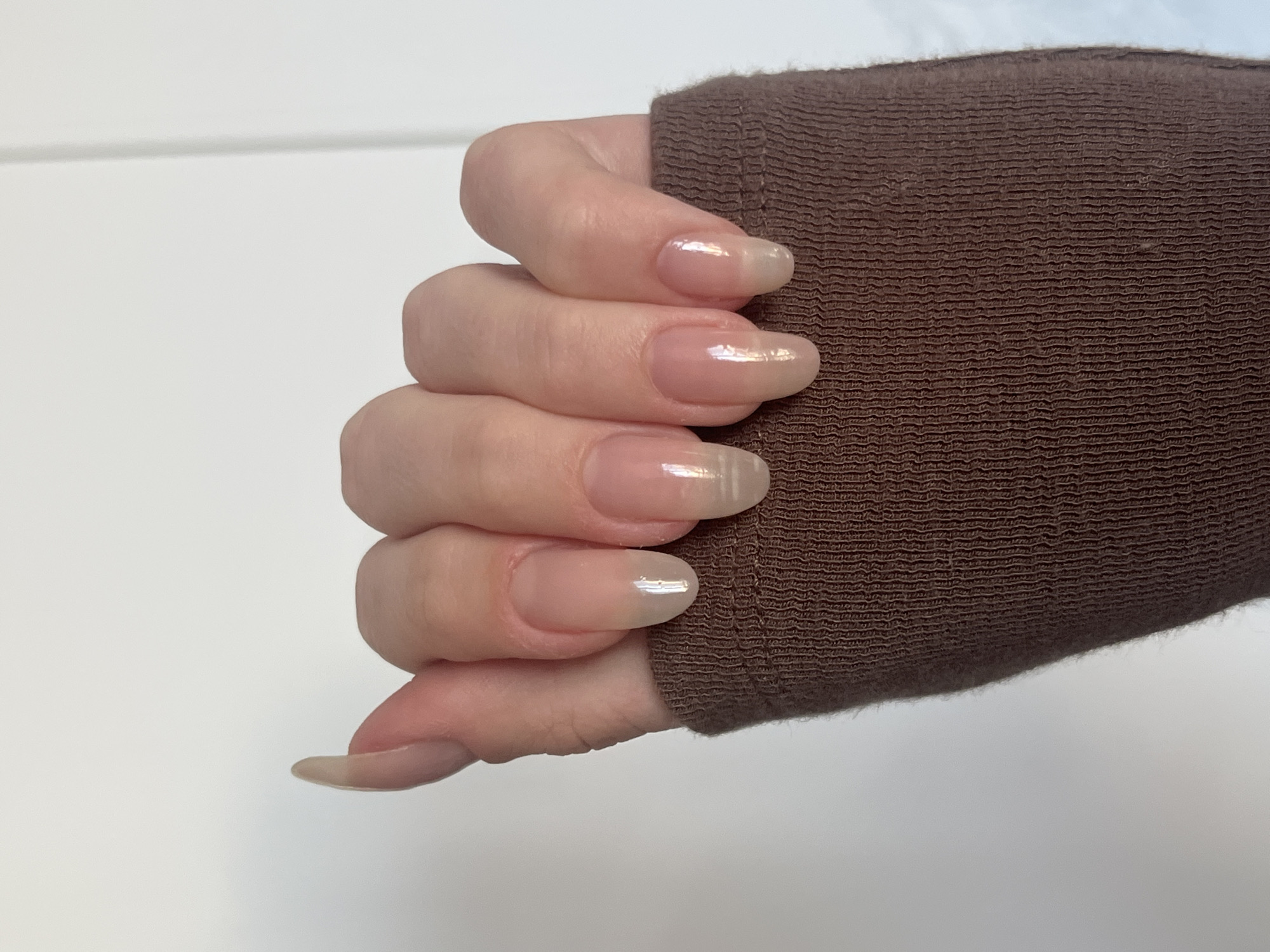 The image displays a close-up of a person's left hand, showing five fingernails against a plain white background. The nails are long, almond-shaped, and appear to have a clear or sheer polish with a subtle iridescent sheen. The hand is partially covered by a brown, textured garment, likely a sleeve, at the wrist and forearm. The lighting creates highlights on the nails, emphasizing their smooth surface. There is no identifiable city or location in Chelyabinsk, Russia, within this image.