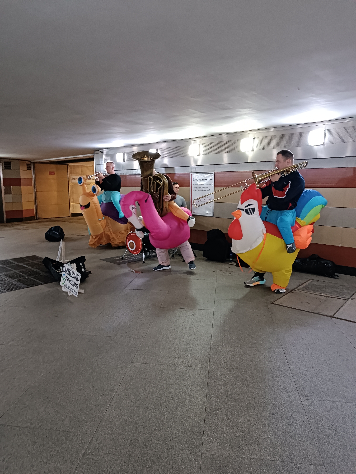 A musical performance is taking place in a subway station. Three individuals, dressed in inflatable costumes of a snail, a flamingo, and a chicken, are playing brass instruments. The snail and flamingo costumes are yellow and pink, respectively. The person in the chicken costume is riding it. A tuba is also present. A drum is set up to the side. A sign for "DUCK BAND" with phone numbers is visible on the ground. The scene is illuminated by overhead lights in a tunnel-like environment with tiled walls. The ground is paved with large square tiles. The image is taken from a medium distance, showing the full figures of the performers and their instruments.