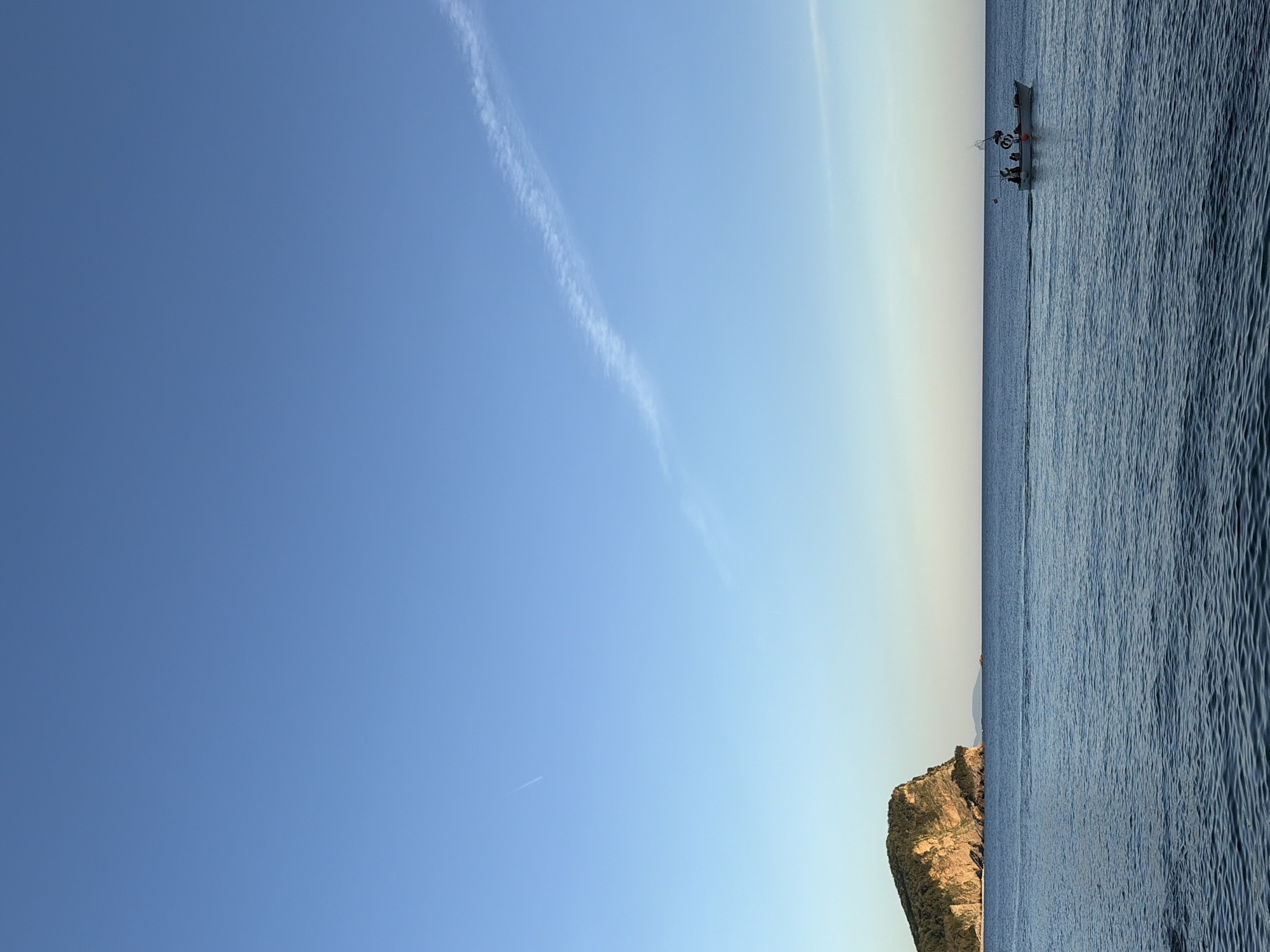 This image, rotated 90 degrees clockwise from its natural orientation, depicts a serene coastal scene under a clear sky. The setting is likely Budva, Montenegro, given the context.
The upper portion of the corrected image features a vast, gradient blue sky, transitioning from a deeper blue at the top to a lighter, almost pale azure near the horizon. A prominent, long, white contrail stretches diagonally across the sky from the top left towards the middle right, indicating recent aircraft activity. Another fainter contrail or distant aircraft is visible further to the right.
The lower half of the image is dominated by the calm, slightly rippled surface of the sea, reflecting the ambient light. On the right side of the sea, a small motorboat or fishing vessel is visible. Several indistinct figures, presumably people, can be seen on board, suggesting a leisurely activity or journey. A dark line, possibly a rope or anchor line, extends from the boat towards the left.
On the left side of the scene, a rugged, rocky cliff or small island rises from the water, displaying shades of brown and green vegetation, suggesting a typical Mediterranean or Adriatic landscape. The light on the cliff face indicates a low sun angle, hinting at either early morning or late afternoon/early evening. The overall atmosphere is peaceful and calm, with mild weather conditions evident from the clear sky and still water. No visible text is present in the image.