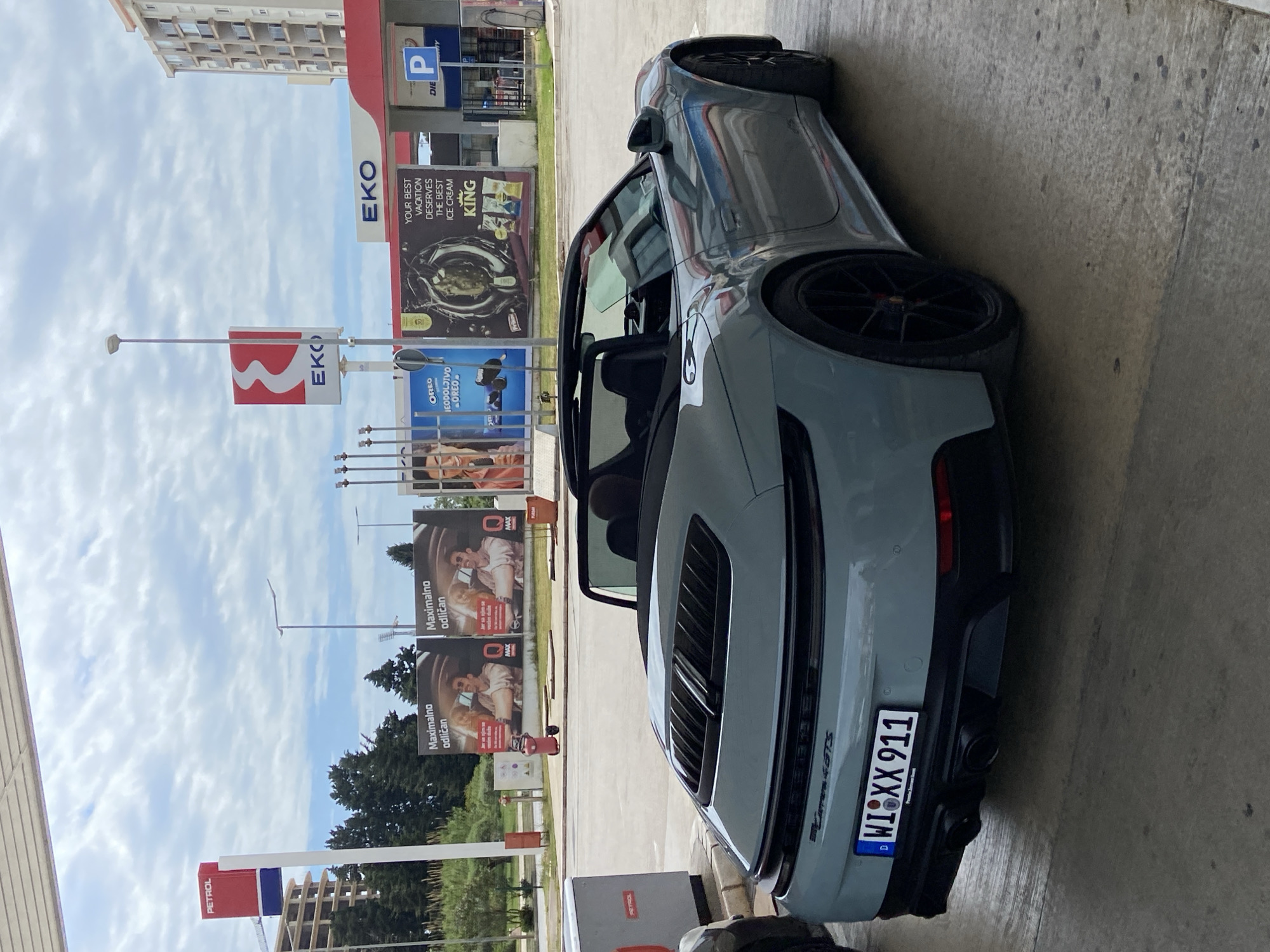 The image shows a gray Porsche 911 Targa 4 GTS convertible parked at a petrol station in Bar, Montenegro. The car has a German license plate "WIXX 911". In the background, there are signs for "Petrol" and "EKO" gas stations. Advertisements for "Oreo" and "King" ice cream are also visible, alongside an advertisement with the phrase "Maximalno odličan". The weather appears to be partly cloudy.