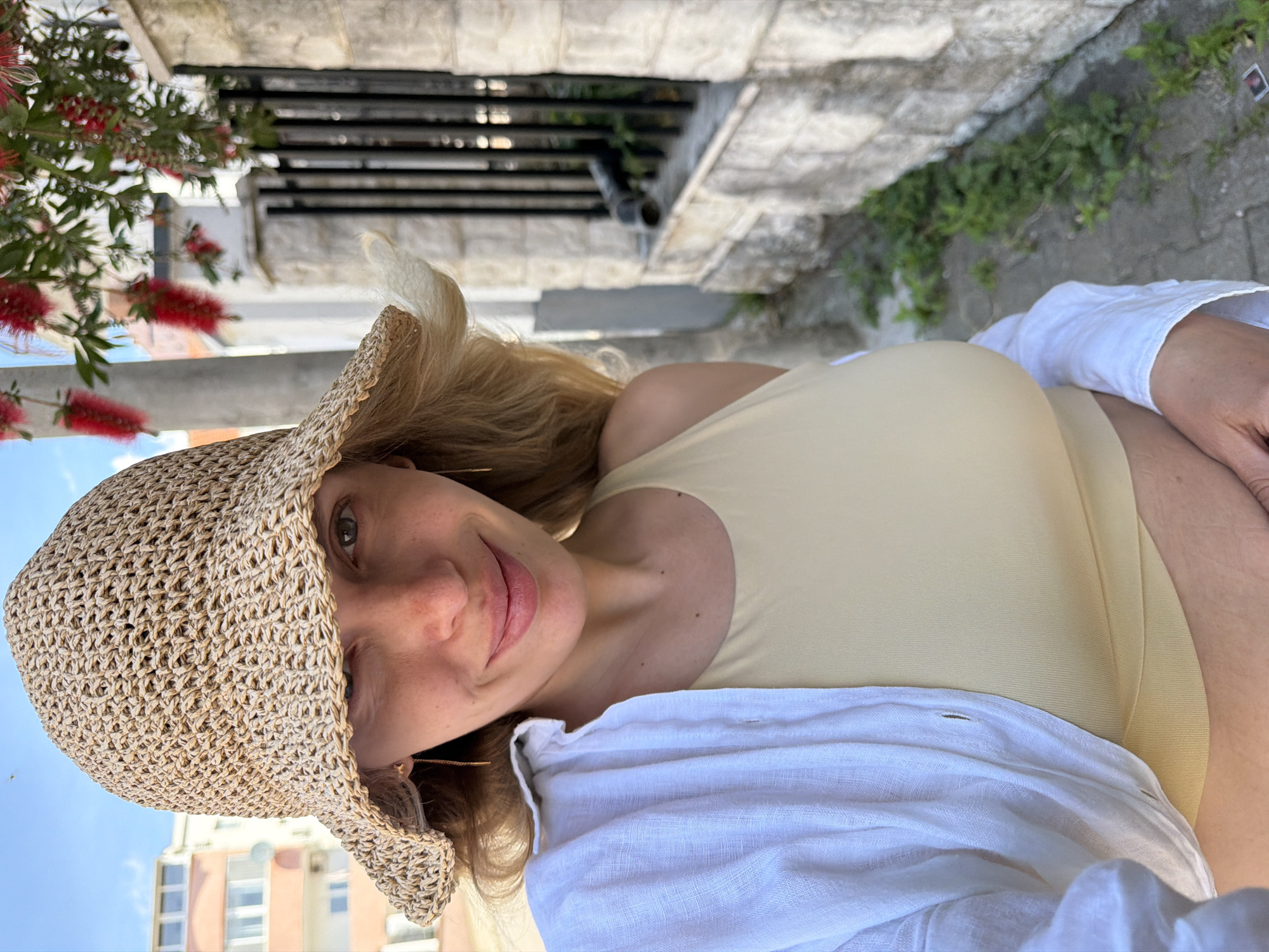 The image features a woman posing outdoors under bright daylight, likely in Budva, Montenegro, as per the provided context. She is seen from the chest up, with a slight tilt to her head. She has long, blonde hair and is wearing a woven straw hat that partially covers her forehead. Her face is directed towards the camera, displaying a soft, pleasant smile. She wears a light yellow or beige-colored tank top or swimsuit, over which a white, loosely worn long-sleeved shirt is draped.
In the foreground to the left, vibrant red bottlebrush flowers with green foliage are prominently visible. The background elements include a rough stone wall or paved surface, suggesting an urban or semi-urban setting. Further back, black metal railings or bars are seen, and a multi-story building with windows is partially visible under a clear blue sky. The overall lighting indicates a sunny day. No visible text is present in the image. The scene conveys a relaxed and pleasant atmosphere.