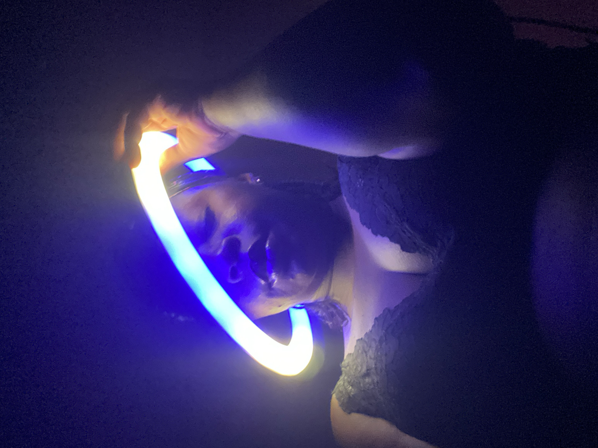 In a dimly lit environment, an individual holds a glowing light stick bent into a U-shape, illuminating their face. The person's features, including their nose and part of their eyes, are visible due to the light source. They are wearing a lace garment that is partially visible in the dark. The location is not identifiable as Maigizo, Nigeria, without further visual cues or contextual information.