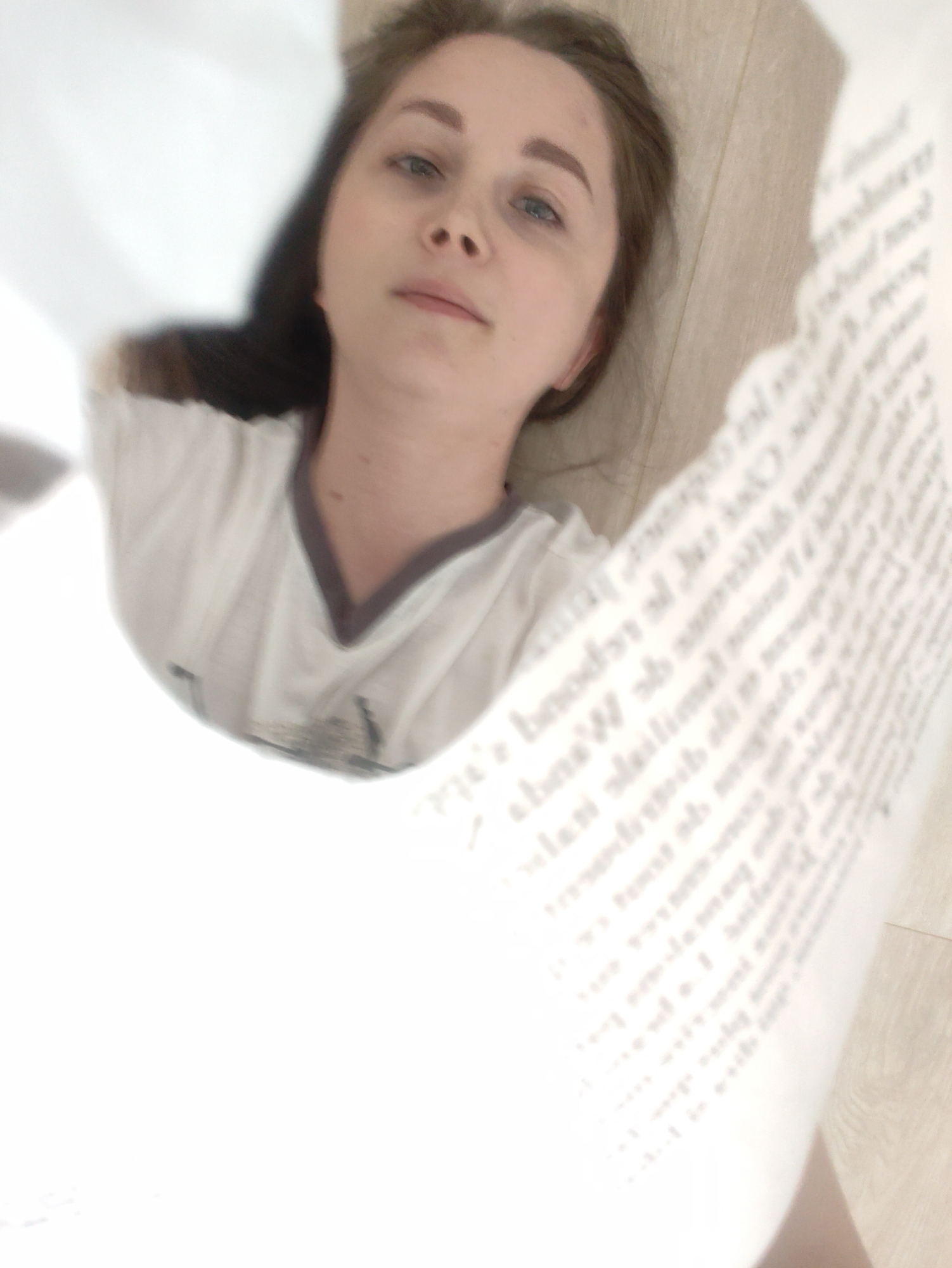 A young woman with light brown hair is viewed through a hole cut in a sheet of paper covered in text. The paper is held close to the camera, partially obscuring the view. The woman wears a white t-shirt with a dark trim around the neck. She is lying on a light-colored wood floor. The location is Unknown, Unknown.