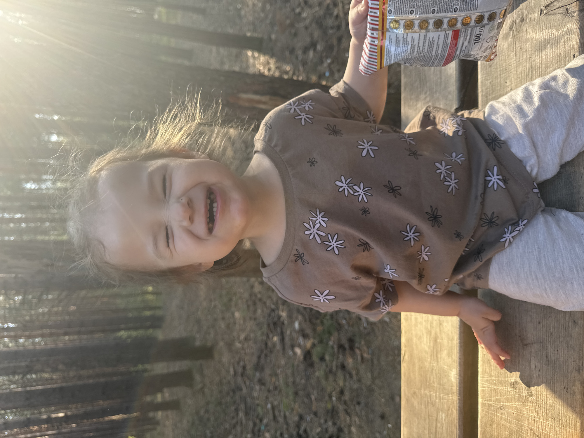 A young child sits on a wooden bench in a sun-dappled area, possibly a park or wooded area in the city of Chaikovsky, Russia. The child wears a brown long-sleeved shirt adorned with white flower patterns and grey pants. The child holds a package of food in one hand and smiles broadly, displaying teeth. The background shows trees and fallen leaves.