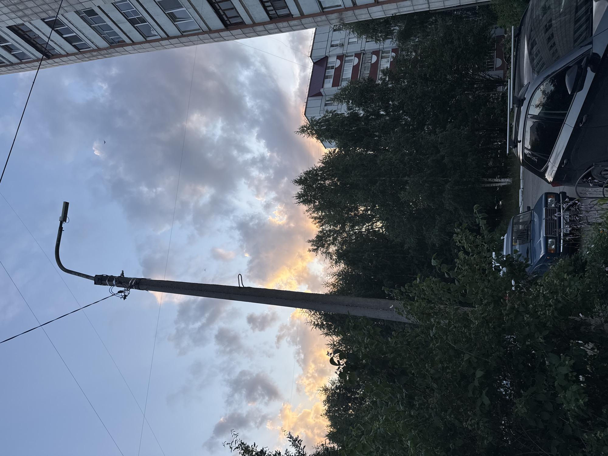 In Chaikovsky, Russia, an outdoor shot captures a twilight scene with a concrete lamppost in the foreground, diagonally bisecting the image. The lamppost is equipped with a street light fixture and overhead wires. Behind the lamppost, a dense line of dark green trees forms a backdrop. In the upper portion of the image, the sky is filled with dramatic, colorful clouds, tinged with orange and yellow hues from the setting sun, contrasting with the cooler blues and grays. To the right of the trees, a multi-story apartment building with a light-colored facade and visible windows stands prominently. Further to the right, another, taller apartment building with a similar architectural style is partially visible. At the base of the trees, two cars are parked. The car on the left is a dark blue sedan, and the car on the right is a dark-colored SUV with a roof rack. The overall impression is an urban landscape transitioning from day to night.