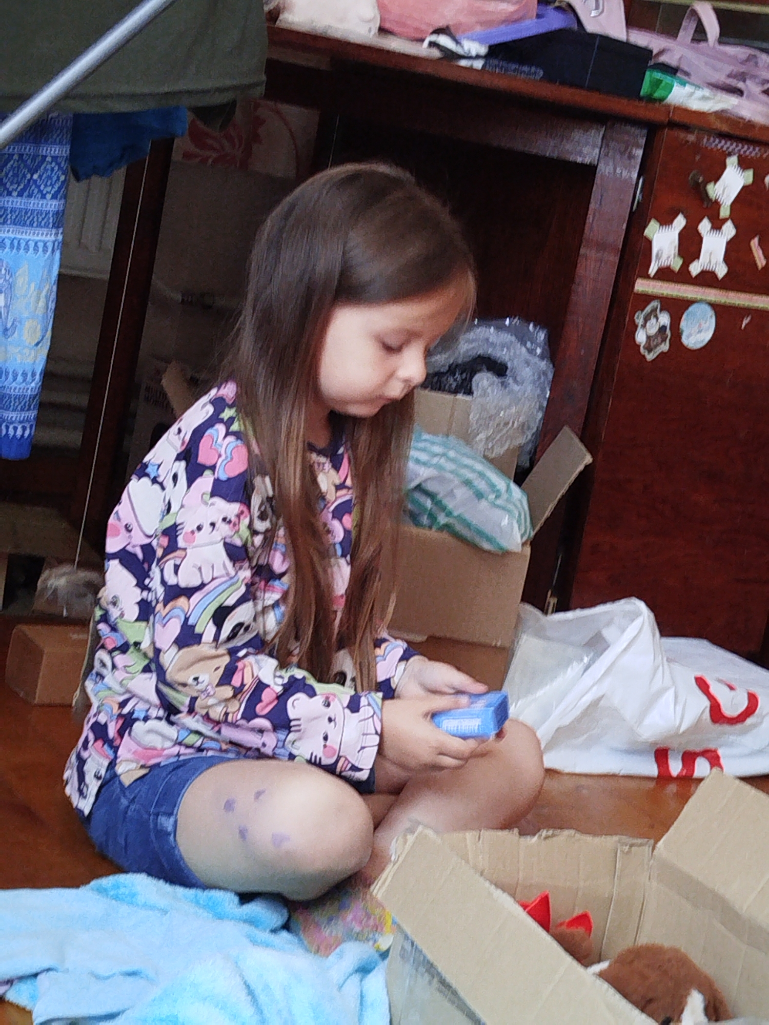 A young girl with long brown hair, wearing a long-sleeved shirt with a pattern of cartoon animals and hearts, and denim shorts, sits on a wooden floor. She is holding a small blue rectangular object in her hands. Her knees are bent, and her legs are visible. To her left, a light blue towel is spread out. Behind her, a cardboard box contains what appears to be a stuffed animal with red accents. To her right, a white plastic bag with red lettering is visible. In the background, a wooden cabinet or dresser is present, with various items on its shelves and decorative stickers on its doors. The lighting suggests an indoor setting. No specific city or location information is discernible from the visual evidence alone.