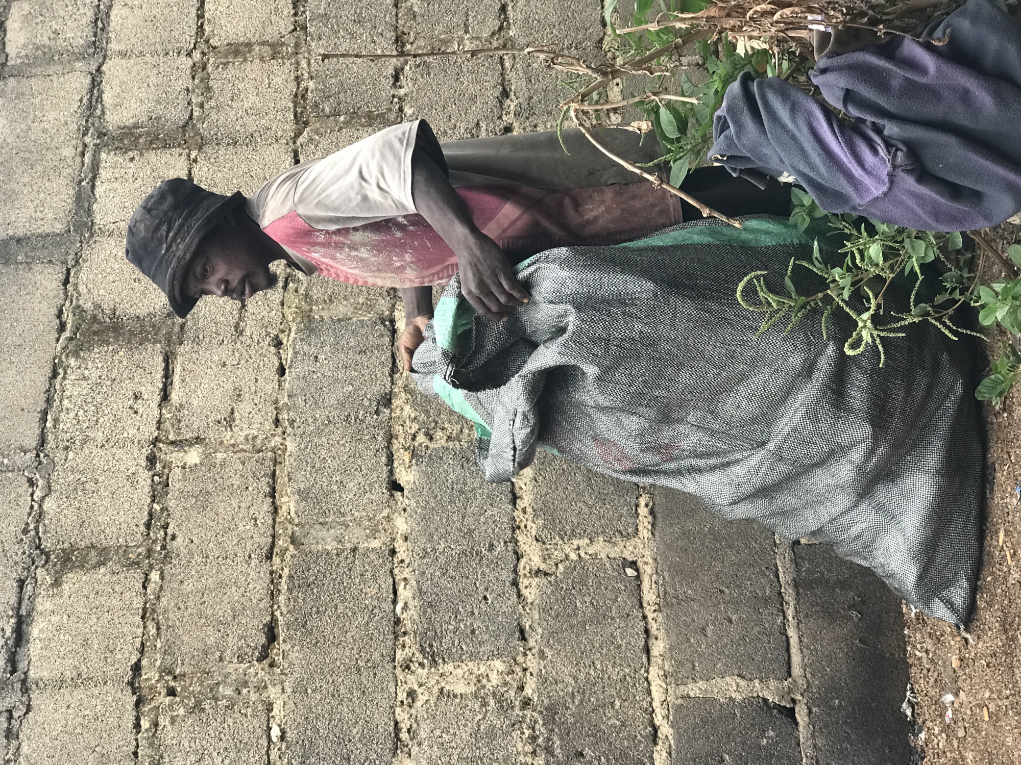 In Kabong, Nigeria, a man wearing a dark bucket hat and a red and grey t-shirt stands next to a large, grey woven sack filled with an unknown substance. The sack is propped upright and appears to be partially open at the top, with the man's hands reaching into it. He is positioned in profile, looking towards the right of the frame, with a slight beard. Behind him, a rough, grey concrete block wall serves as a backdrop, showing signs of weathering. To the right of the sack, some green vegetation and dry branches are visible, along with a crumpled dark blue and purple cloth. The ground is covered with loose dirt and small debris.
