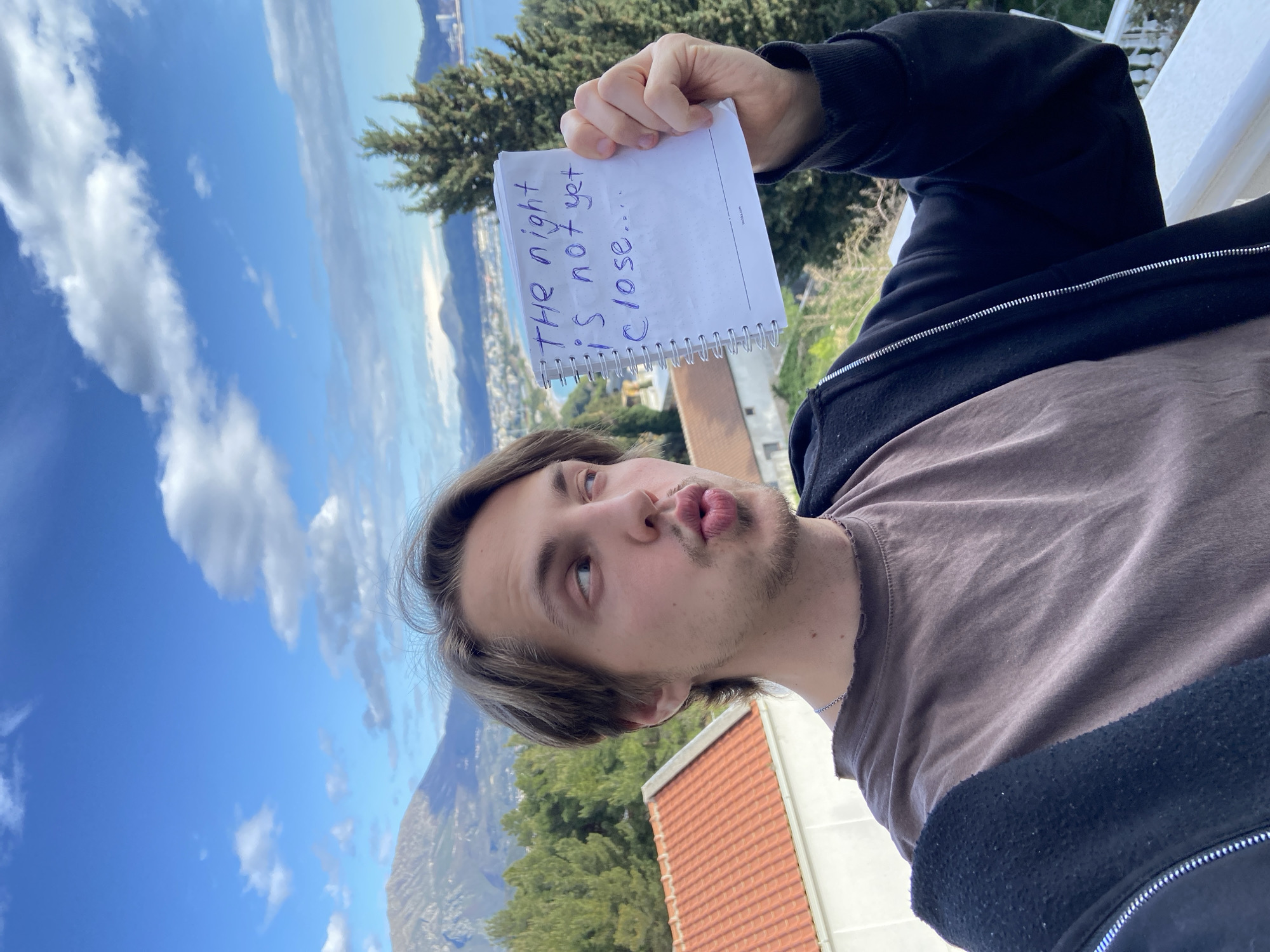 A male subject with light brown hair and a slight beard holds a spiral-bound notebook with the handwritten phrase "THE night IS not yet close..." in blue ink. He is wearing a brown t-shirt and a black hooded jacket. He is looking upwards and pursing his lips.
In the background is a view of the coastal city of Šušanj, Montenegro, with buildings, greenery, and mountains visible. The sky is blue with scattered white clouds. A tree is visible behind the notebook, and what appears to be a red-tiled roof is in the foreground.