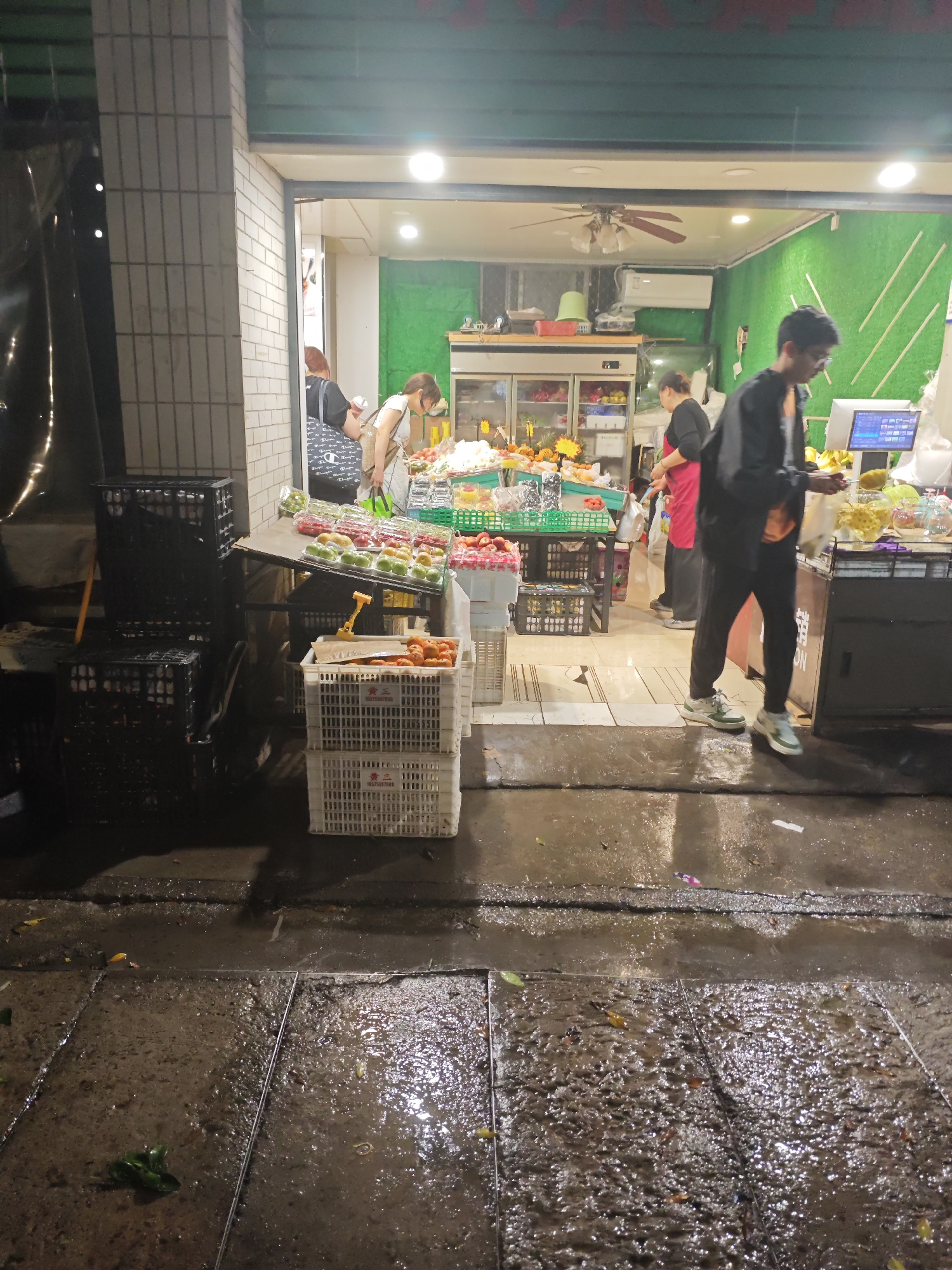 The image depicts a wet, potentially rainy, street scene outside a fruit and vegetable store in Chongqing High-tech Zone, China. The store's entrance is brightly lit, showcasing a variety of produce arranged on tables and within a refrigerated display case. Several people are present inside the store: a woman in a white shirt appears to be shopping, another woman in a black shirt and red apron stands behind the counter, and a man in a black jacket stands outside near the payment area. Outside the store, there are several crates of produce and empty black plastic crates stacked beside the store's entrance. The street is wet and reflects the store's light, with some leaves and debris scattered across the surface. The store is built into a building structure of light-colored bricks.