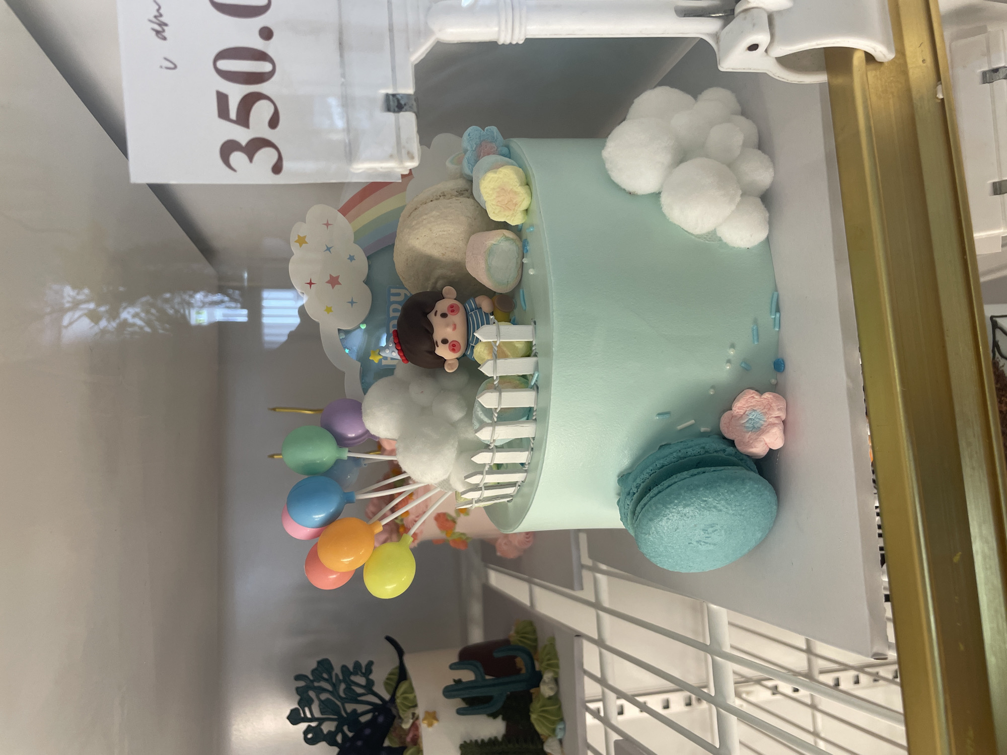 A light blue frosted cake decorated with a cartoon boy figure, miniature white picket fence, marshmallows, and balloons is displayed on a white cake stand. Next to the cake is a blue macaron and a pink flower-shaped candy. In the background, another cake is visible with a cactus and dinosaur decoration. A price tag in the upper right corner reads "350.0". The scene is indoors, likely in a bakery or display case.