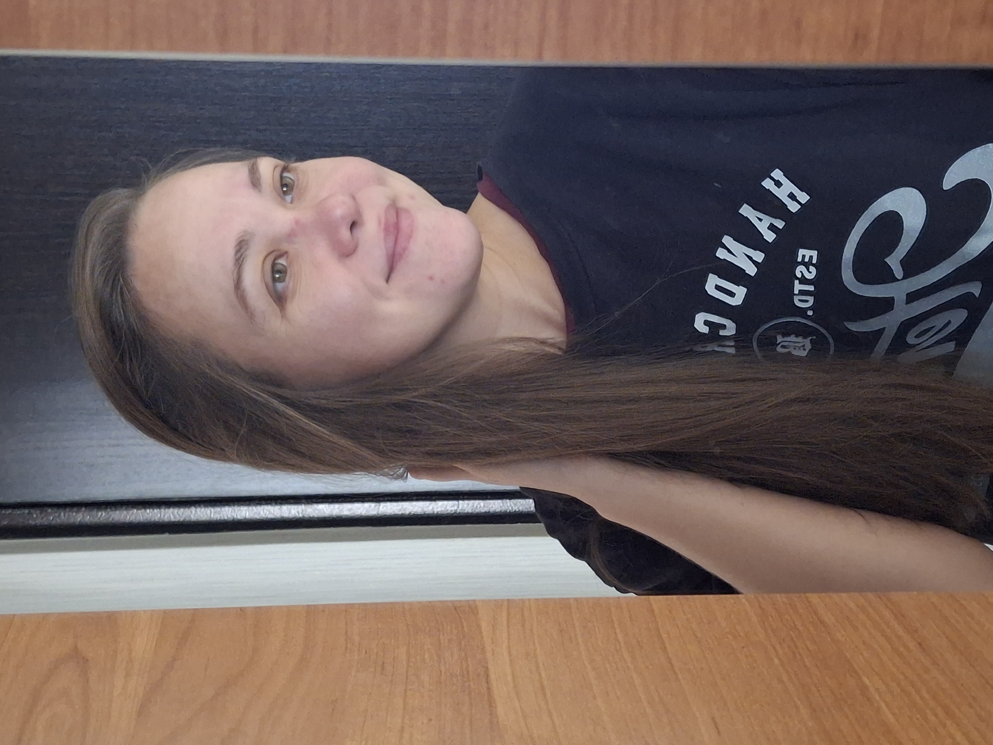 A woman with long brown hair is pictured in a mirror reflection, indoors. She is looking to her right and smiling slightly. She is wearing a dark t-shirt with white text and graphics on it. The background appears to be a doorway or a similar architectural feature, with wood-toned paneling on the left and a dark surface behind her. The image is taken from a medium shot, focusing on her upper body and face. The location is identified as Chaikovsky, Russia.