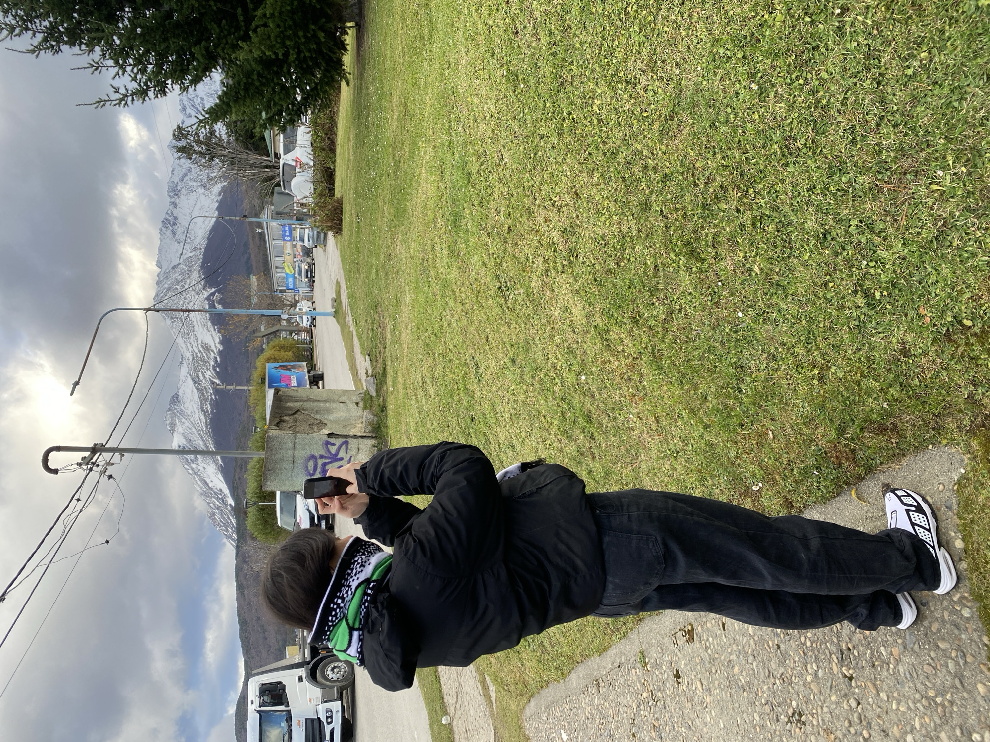 The image shows a person taking a photograph with a smartphone in Ushuaia, Argentina. The individual is standing on a paved sidewalk with a grassy area to their right. They are wearing a black jacket, black pants, and white sneakers with black detailing. A patterned scarf is visible around their neck. In the background are mountains with patches of snow. A street with parked vehicles and utility poles is also visible in the background. The sky is overcast.