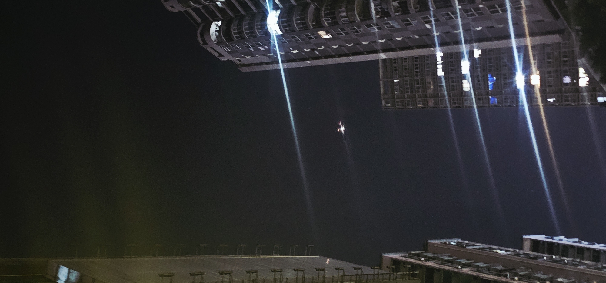 This is a photograph taken at night in the Jiulongpo District, China. The image features the exteriors of several multi-story residential buildings against a dark sky. The perspective is looking upwards, with the buildings dominating the frame. One building on the left, partially visible, is tall and appears to be illuminated by internal lights that create a yellowish glow. In the center of the frame, a smaller object, possibly a drone or a small aircraft, is visible in the sky. It appears to have multiple lights and is leaving a faint trail. On the right side of the image, another tall residential building is prominently displayed. This building has numerous windows, many of which are lit from within, showing various colors of light, including white and blue. Streaks of light, likely lens flares or light reflections from the buildings, are visible crossing the sky diagonally. The overall scene depicts an urban nightscape with a focus on the architecture and the sky.