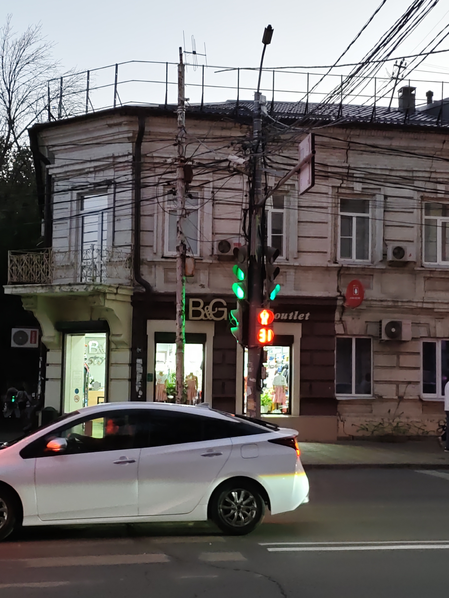 The image captures a street scene in Rostov-on-Don, Russia, on a cloudy day. In the foreground, a white car, likely a hybrid model given its sleek design and the visible Toyota logo on the rear, is driving on a paved road. The car is positioned on the right side of the road, suggesting it is being driven in accordance with Russian traffic laws. The car's headlights are on, illuminating the road ahead and casting a subtle glow on the asphalt.
Dominating the background is a multi-story building with an aged facade, characterized by ornate architectural details, balconies, and numerous electrical wires tangled around a utility pole. The building appears to house several businesses on the ground floor, indicated by illuminated storefronts. One storefront displays the "B&G outlet" signage, with mannequins dressed in clothing visible through the window. Another shop also appears to be a clothing store, with mannequins and clothing racks inside. The architecture of the building suggests it might be a historical structure.
A traffic light is prominently visible, showing red lights for vehicle traffic. The sky above is overcast and a pale grey, typical of a cloudy day. The overall impression is of a bustling urban environment, with a mix of modern vehicles and older buildings, characteristic of many cities in Russia. The presence of the car, the traffic light, and the shops indicate ongoing commercial and vehicular activity.