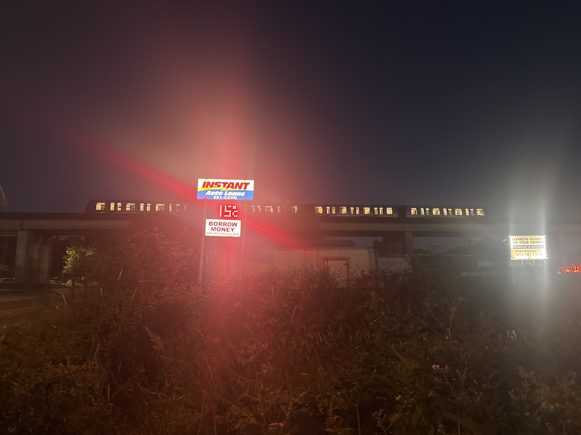 The image depicts an exterior scene at night in Surrey, Canada. A train is seen on an elevated track passing by a lit-up billboard for "INSTANT Auto Loans 581-CASH." The billboard also displays the number "152" in red illuminated digits. To the right, another billboard advertises "BORROW MONEY ON YOUR VEHICLE," with "LOANS IN 15 MINUTES" and the website "INSTANTAUTOLOANS.COM 604 581-2274." The foreground is filled with dense vegetation and tall grasses, illuminated by the glowing signs. The overall atmosphere is dark with strong artificial light sources from the billboards and the train.