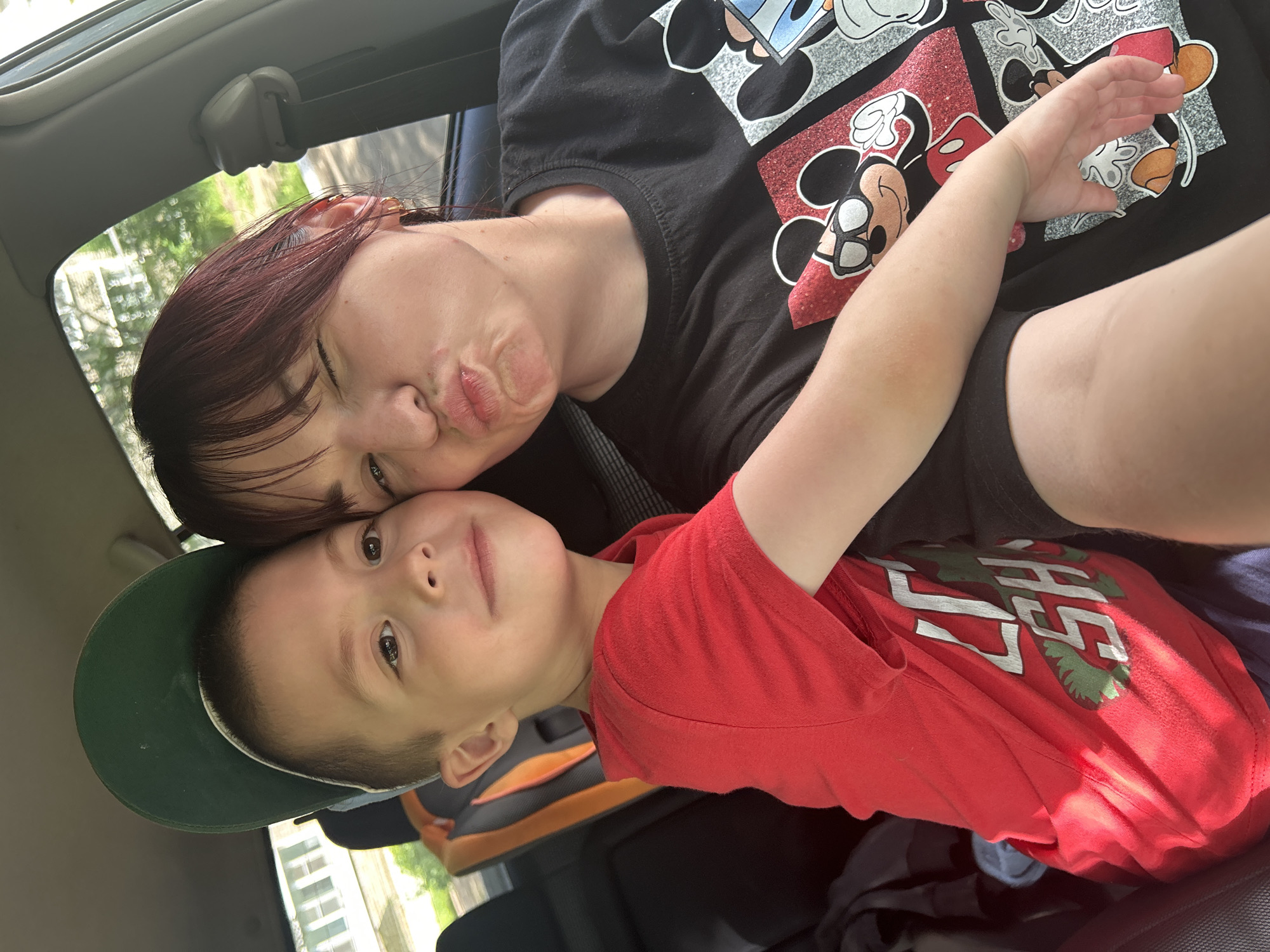 Two people, a young boy and an adult female, are in a car. The boy is wearing a red t-shirt and a green baseball cap backward. He is leaning against the female, who is wearing a black t-shirt with Mickey Mouse graphics. The female is making a kissing face and winking. They are both looking at the camera. The interior of the car is visible, including car seats and a seatbelt. Outside the car, trees and buildings can be seen through the windows. The lighting suggests it is daytime. The scene appears to be in a residential area. The location in the city of Chaikovsky, Russia, is not discernible from the image.
