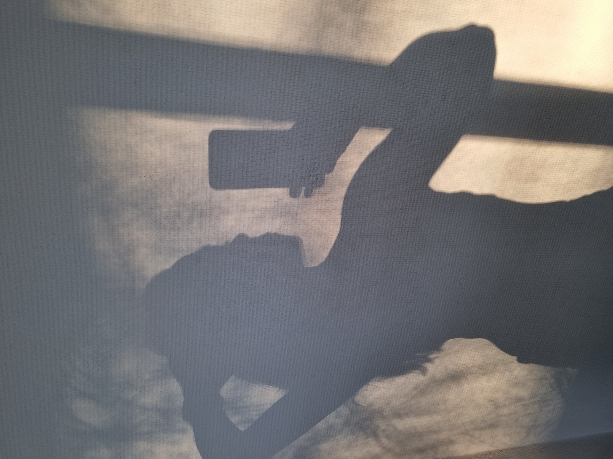 The image depicts the shadow of a person holding a smartphone, seemingly taking a picture. The shadow is projected onto a textured surface, possibly a curtain or wall. The scene appears to be indoors. There is no information in the provided image that indicates the location is in the city of Kemul, Russia.