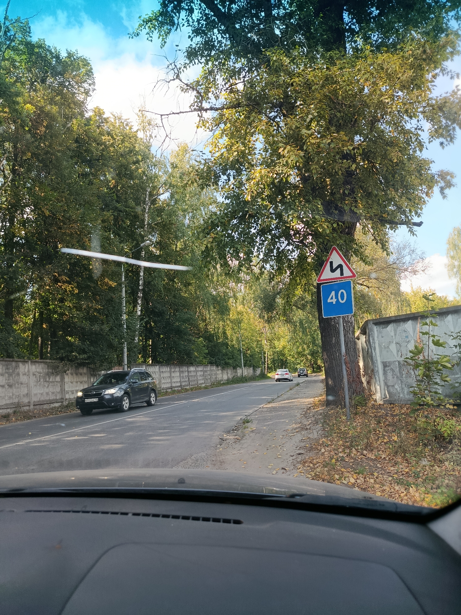 The image depicts a view from inside a vehicle, looking out at a road in Orekhovo-Zuyevo, Russia. The road is paved and appears to be a two-lane street. On the right side of the road, there is a large tree with a road sign attached to its trunk. The sign is triangular and shows a zigzag symbol, indicating a winding road, and below it is a blue rectangular sign with the number "40" in white, signifying a speed limit of 40 kilometers per hour. A grey concrete fence runs along the left side of the road, separating it from a grassy area and more trees. In the distance, other vehicles are visible on the road, including a dark SUV in the foreground on the left and two lighter-colored cars further down. The environment is characterized by lush green trees, suggesting it is either late spring or summer, with a clear blue sky and scattered clouds visible above. Fallen leaves are scattered on the ground near the road and the fence, hinting at the approach of autumn. The overall scene is a typical roadside view in a semi-urban or suburban area.