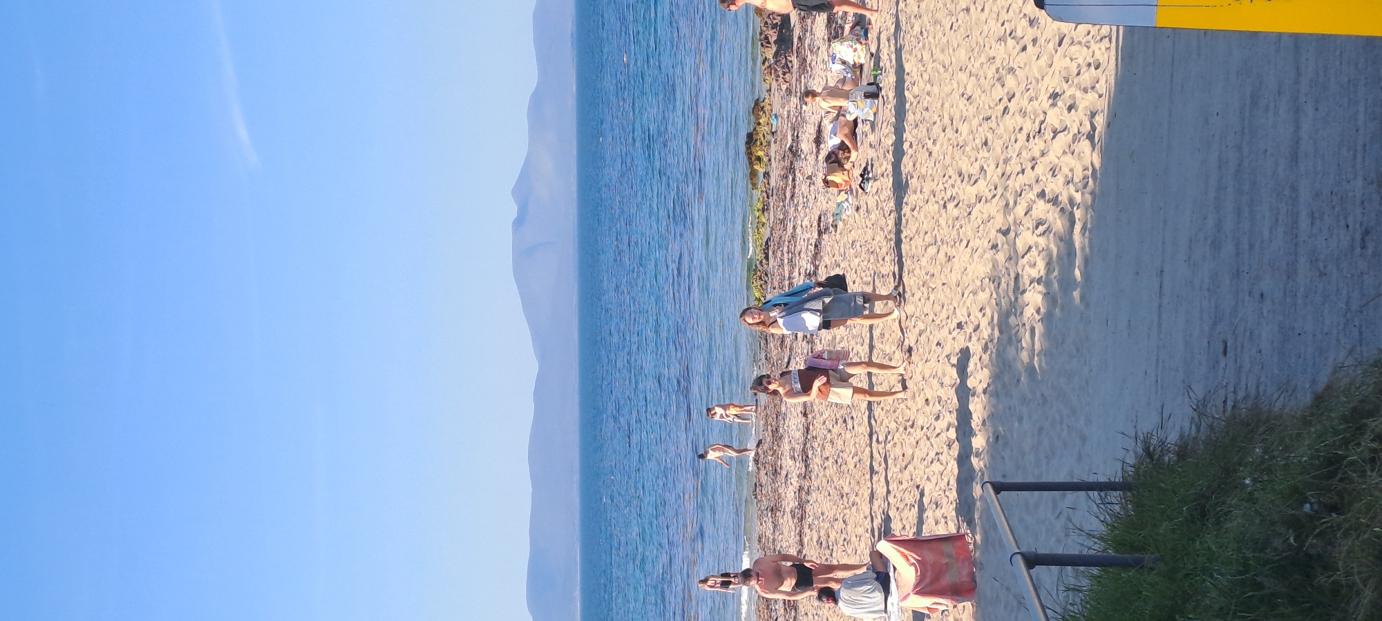 This is a vertical, eye-level shot of a beach scene on a sunny day, looking out towards the sea and a distant mountain range. The sand is light brown and textured with ripples. In the foreground, a metal railing with two vertical posts borders a patch of green grass. The sea is a vibrant blue, with small waves lapping at the shore. Several people are scattered across the beach and in the shallow water.
In the mid-ground, there are two women walking on the sand. The woman on the left is wearing a brown and white patterned top and light-colored shorts, carrying a pink bag. The woman on the right is wearing a blue jacket over dark clothing and carrying a grey towel and a dark bag. To their left, a man in black swim trunks stands on the sand, facing the water. Further to the left, a person in a light-colored shirt and dark shorts sits on a red towel. In the water, several people are visible, some wading and others further out.
In the background, the sea stretches to the horizon, meeting a hazy, purple mountain range under a clear blue sky. The overall impression is a relaxed, summer day at the beach in Kilsallagh, Ireland.