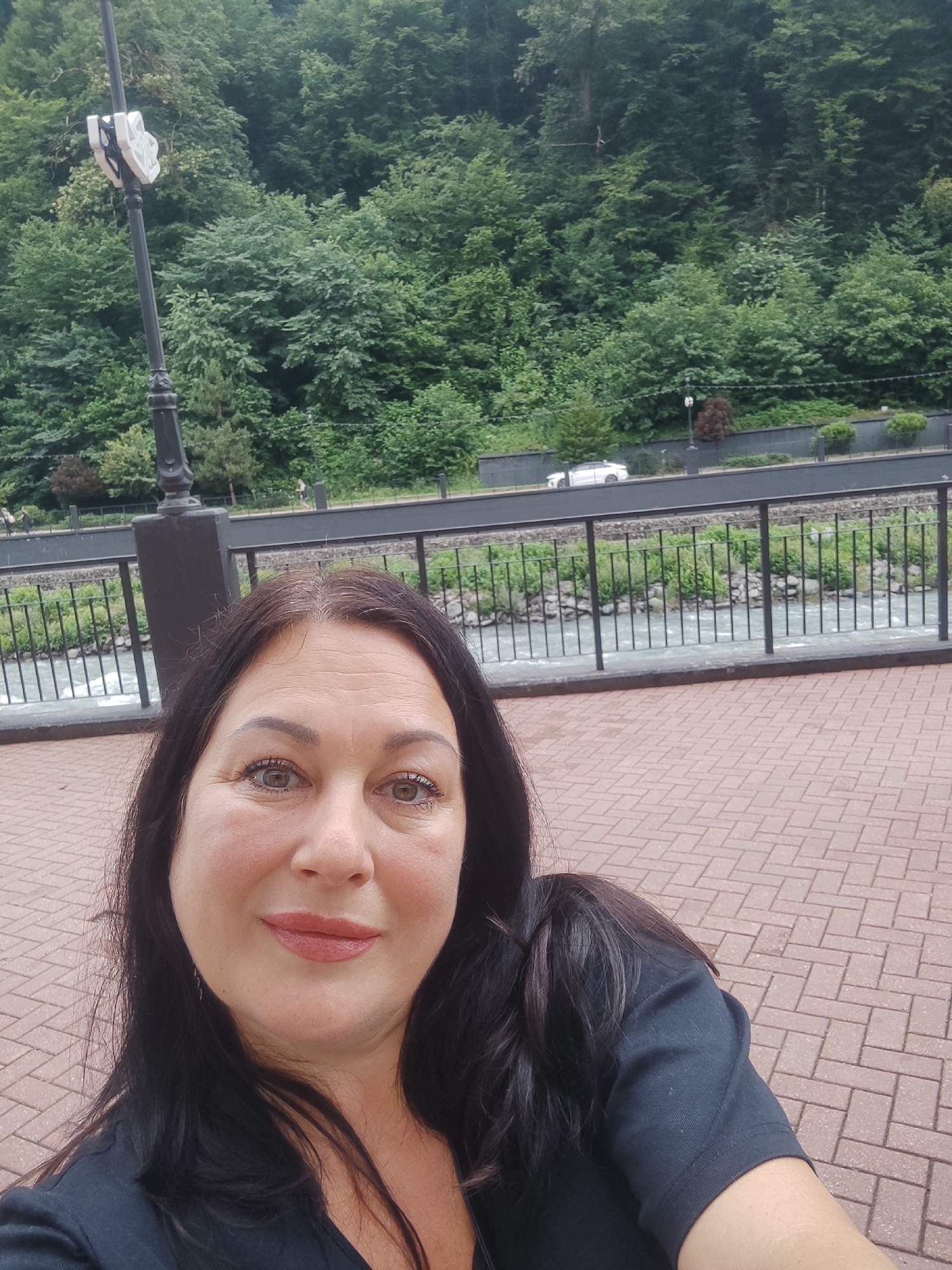 A woman with long dark hair takes a selfie in front of a wooded mountain slope in Estosadok, Russia. She is wearing a black shirt. Behind her, a river flows past a paved walkway bordered by a metal fence. Trees cover the hillside, and a white car is visible on a road in the distance. A streetlamp is attached to a dark pole to the left of the frame. The ground in front of her is paved with brick-like tiles.